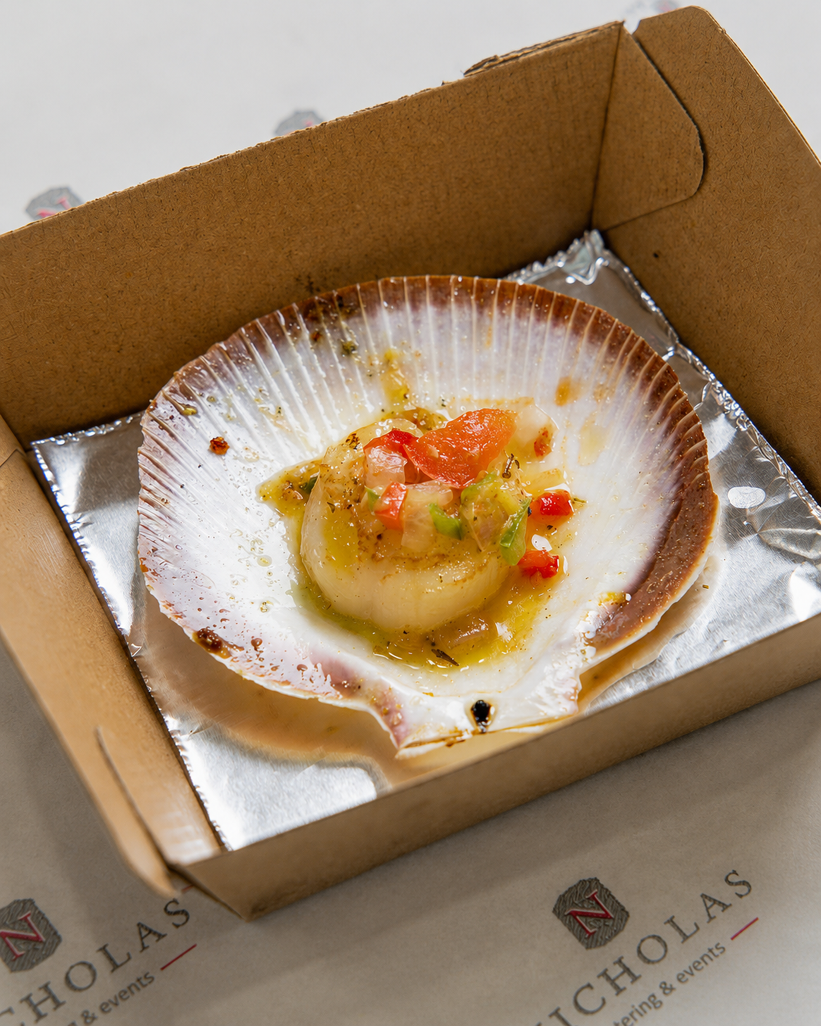 A partially eaten scallop served in its shell with chopped vegetables and sauce, placed in a cardboard takeout container lined with aluminum foil.