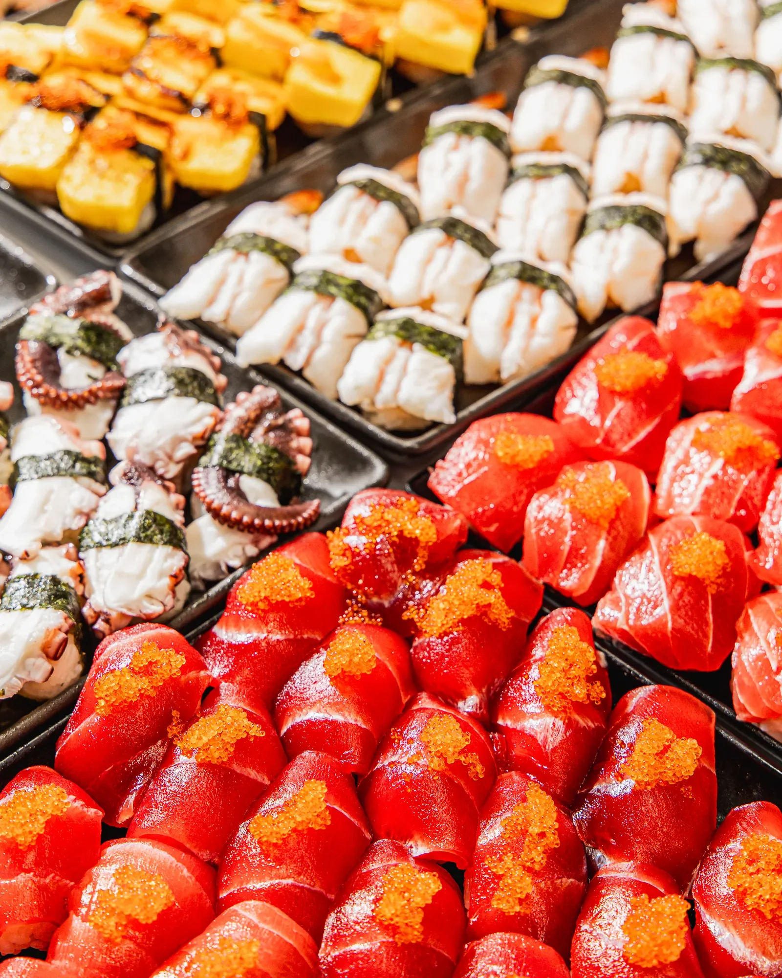 Close-up of assorted sushi rolls and sashimi, including salmon, octopus, and other types, arranged on black trays at a sushi restaurant or buffet.
