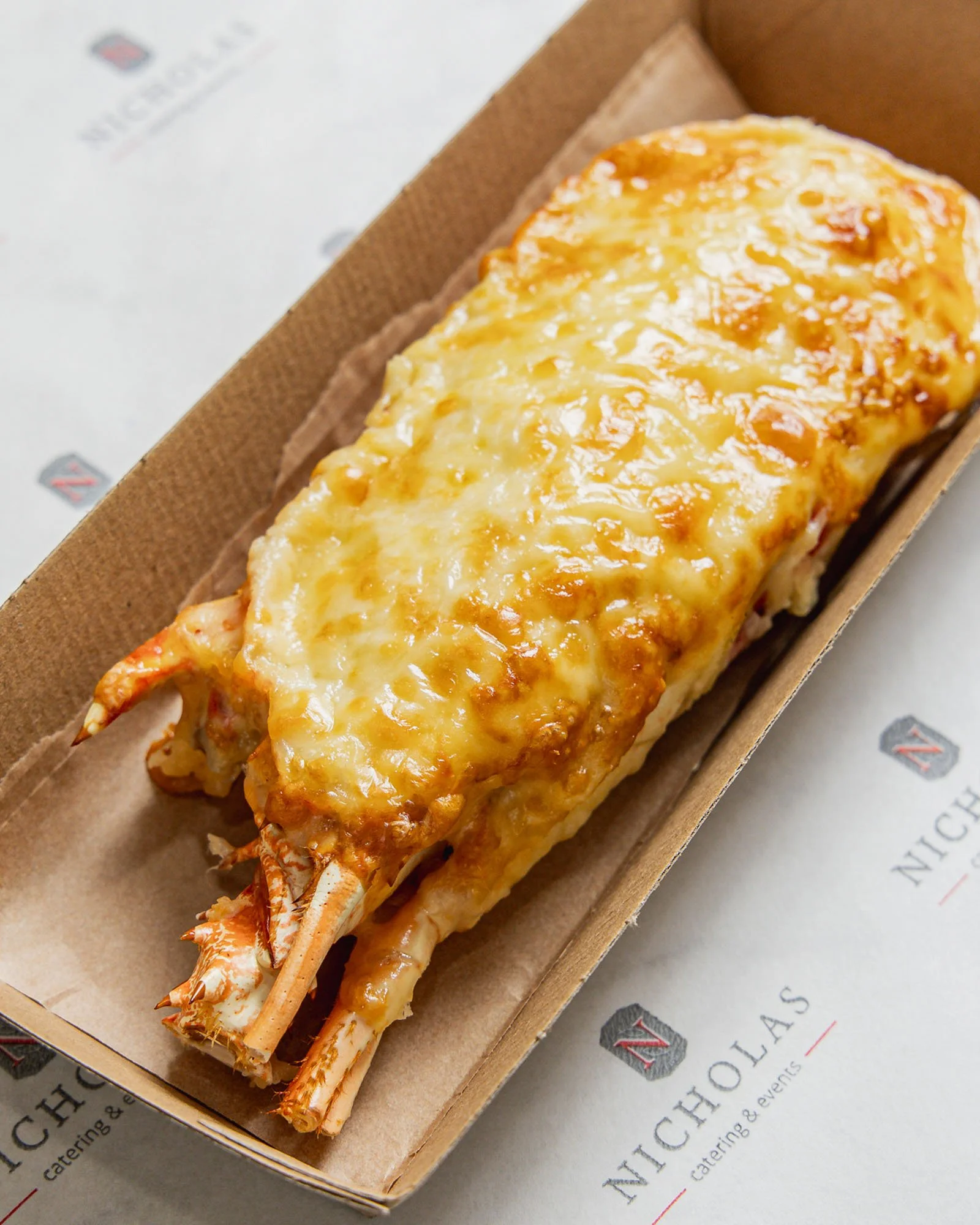 Lobster tail topped with melted cheese, inside a cardboard takeout container.