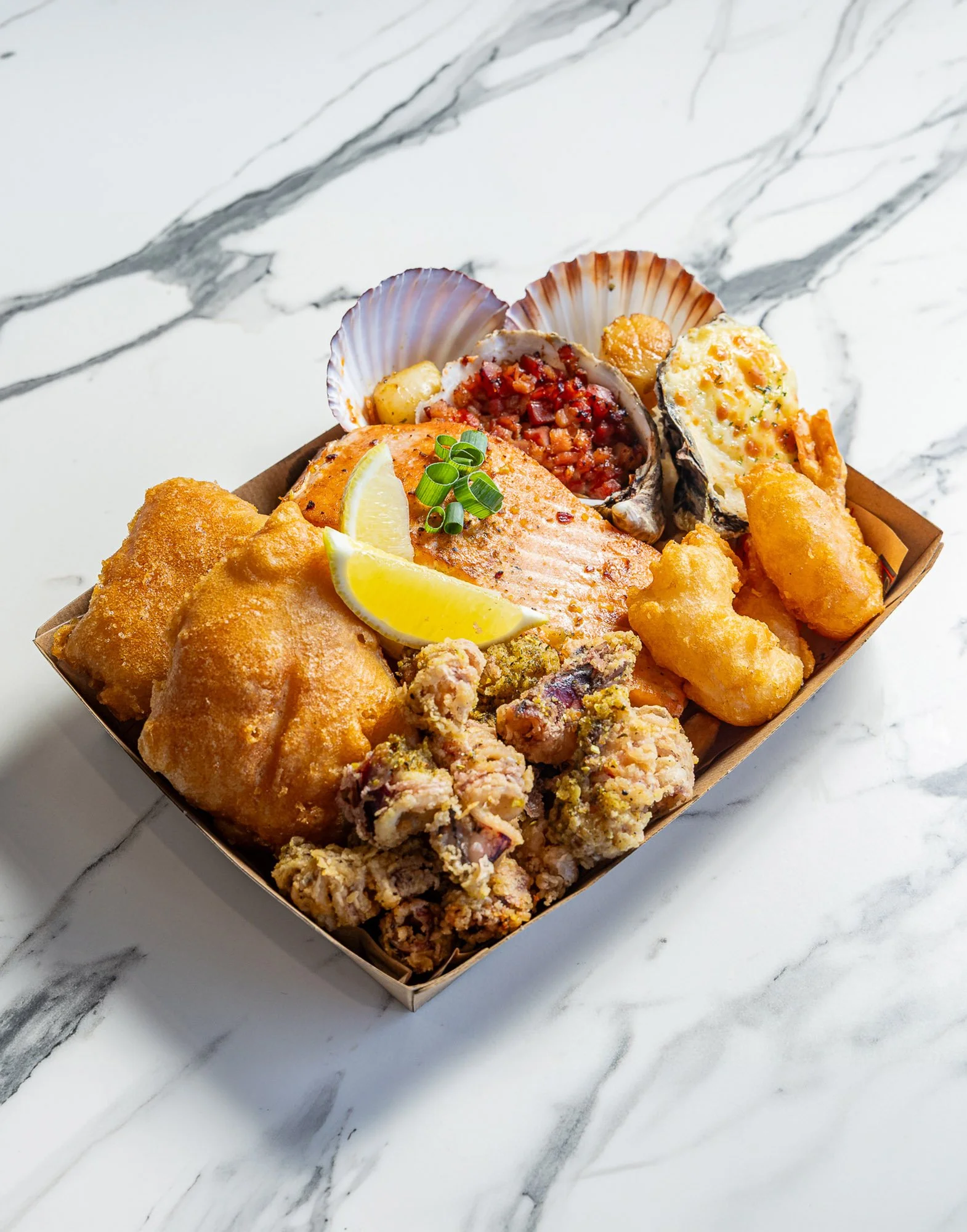 Variety of fried seafood including fish fillets, crab cakes, and stuffed shell, garnished with lemon wedges and green onions, served on a marble countertop.