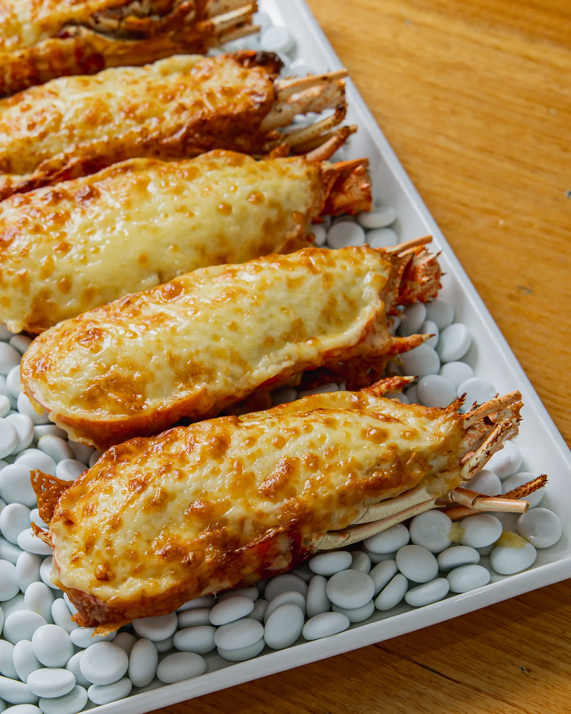 Close-up of lobster tails topped with melted cheese, arranged on a white serving platter with white beans underneath, on a wooden table.