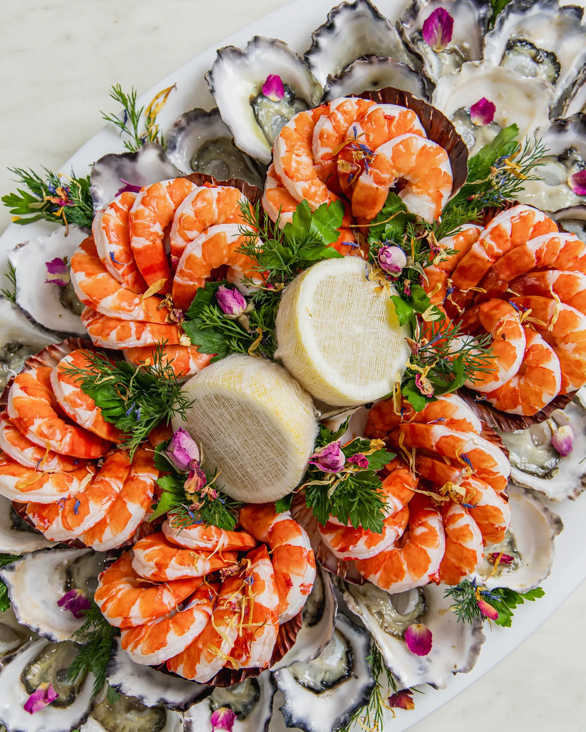 A platter of oysters on the half shell garnished with fresh herbs, purple and pink edible flowers, and topped with shrimp and lemon slices.
