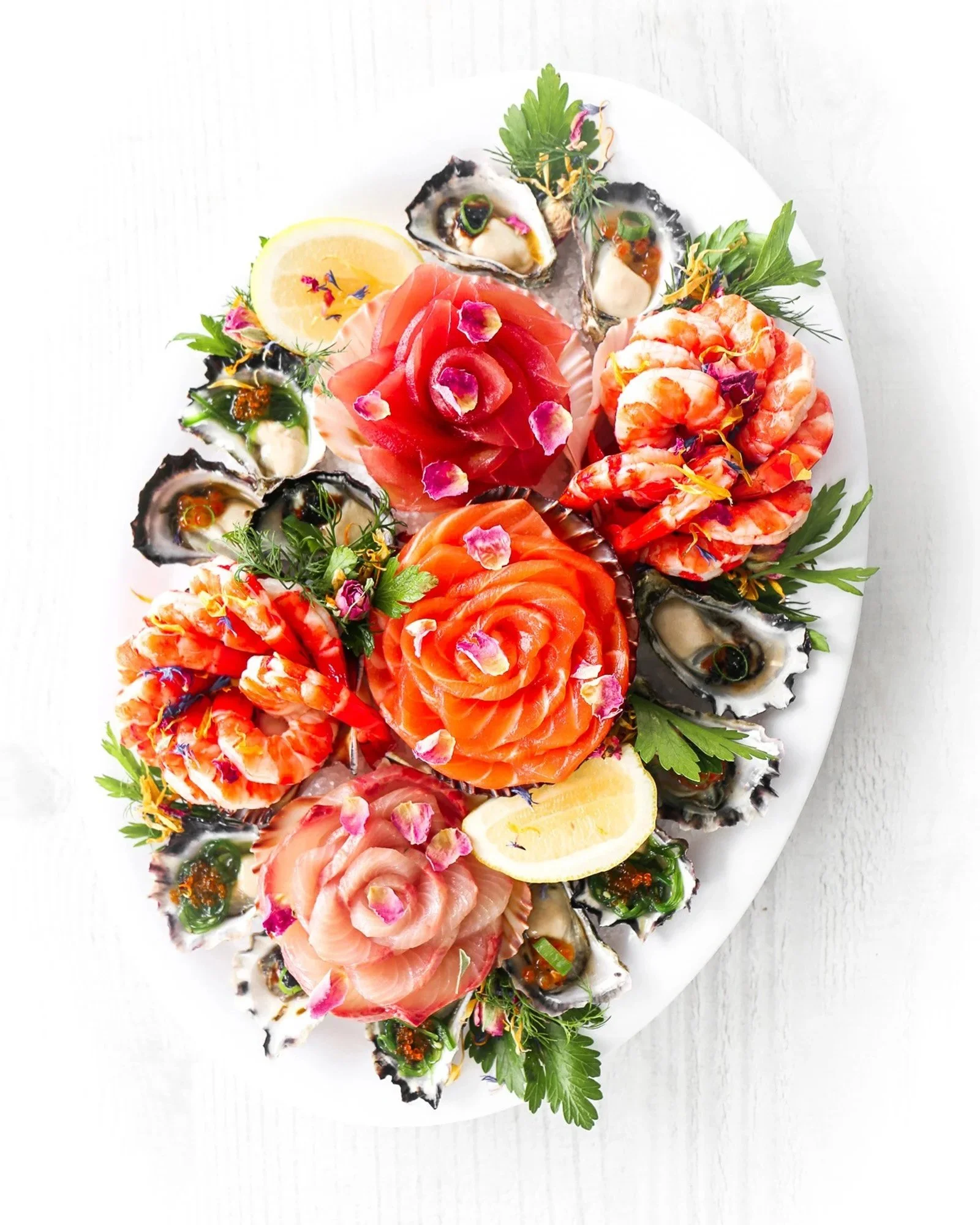 A white oval platter with assorted raw seafood including oysters, shrimp, and slices of salmon, garnished with lemon wedges, fresh herbs, and edible flower petals on a white surface.