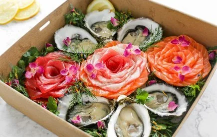 Assorted sashimi and oysters garnished with edible flowers in a cardboard box
