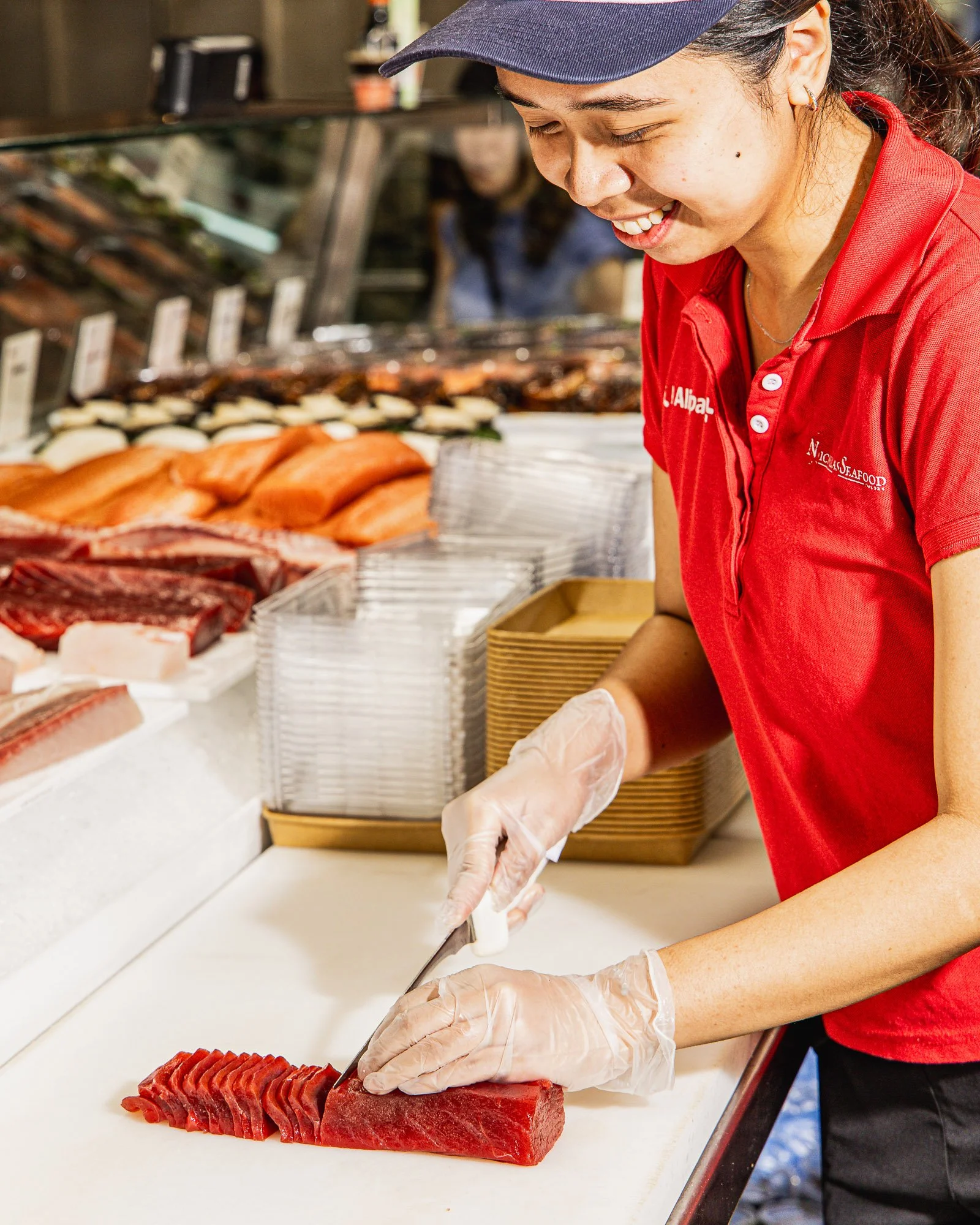 A person wearing gloves and a red shirt preparing sliced raw tuna fish at a sushi or seafood counter.