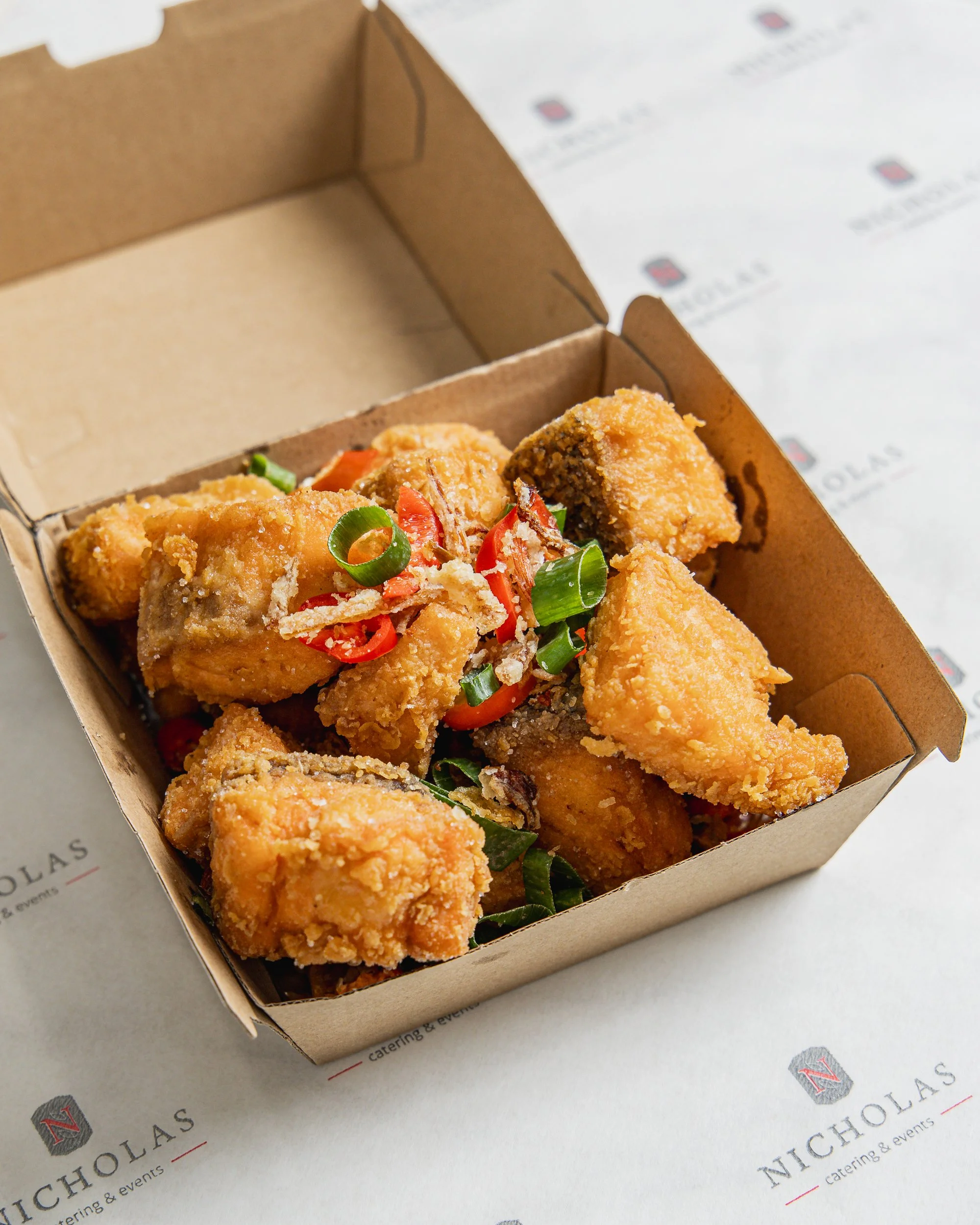 Fried chicken pieces topped with chopped green onions and red chili peppers inside a cardboard takeout box on a branded paper surface.