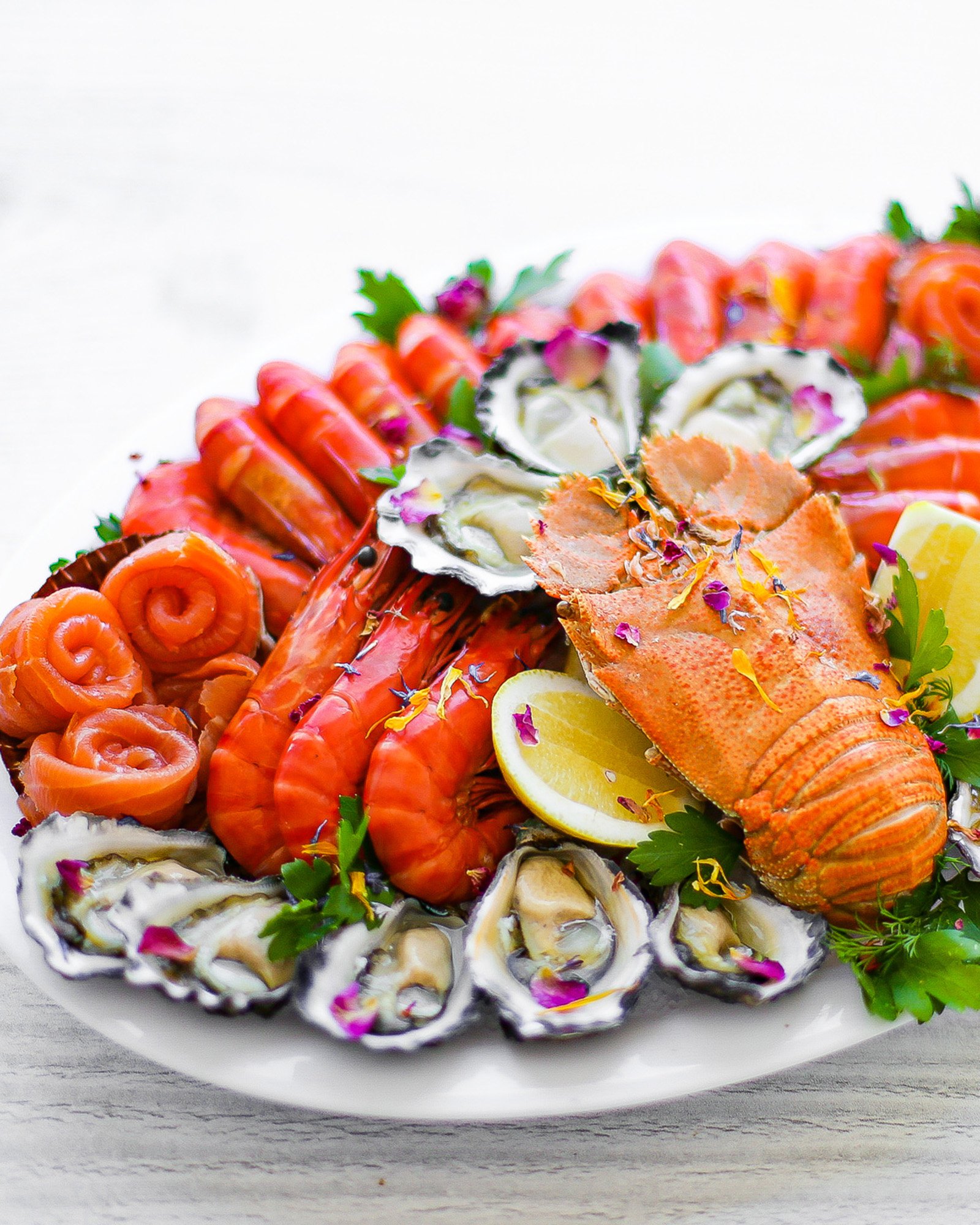 Seafood platter with oysters, smoked salmon rolls, lobster tail, lemon slices, and garnishes on a white plate.