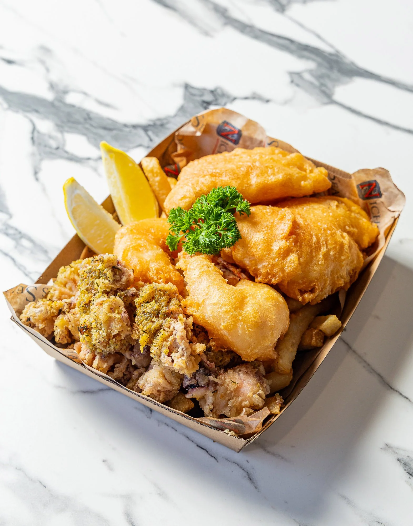 A basket of fried fish fillets garnished with lemon wedges and parsley, with a side of crispy breaded chicken wings, on a white marble surface.