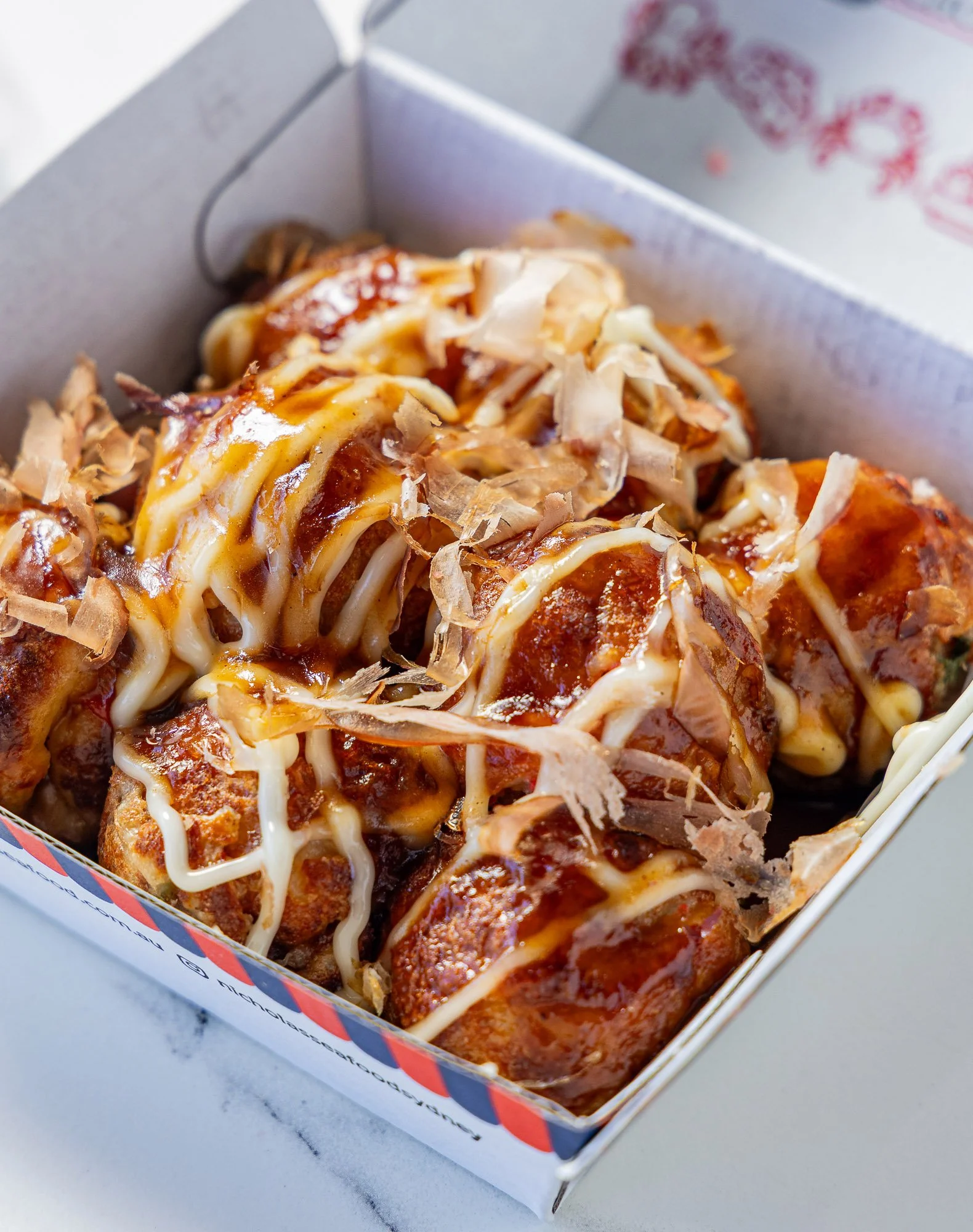 Close-up of takoyaki, Japanese octopus-filled snack balls topped with sauce, mayonnaise, and bonito flakes in a white takeout box.