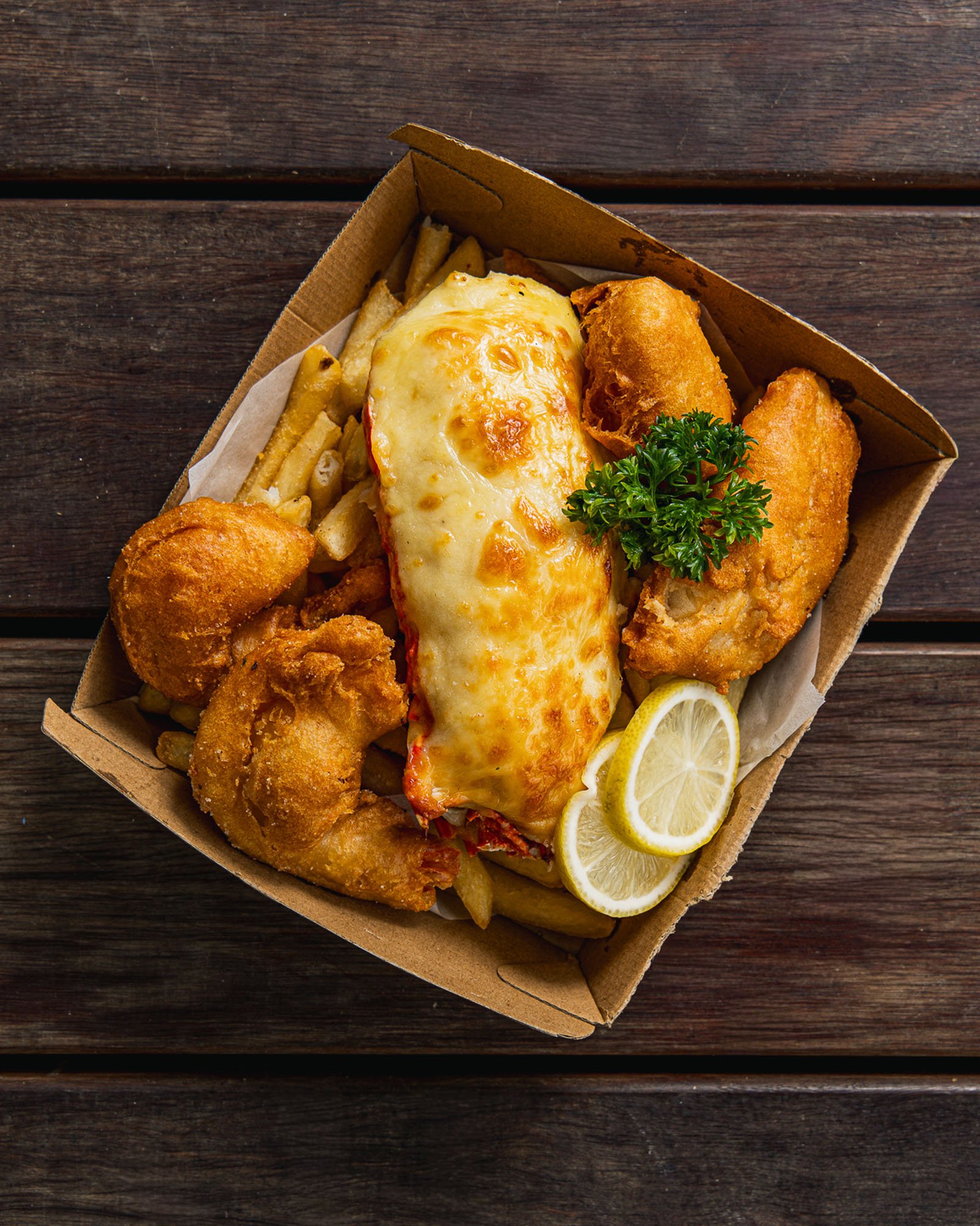 A takeout container filled with fried fish fillets, cheeseburger with melted cheese, French fries, lemon slices, and parsley on a dark wooden surface.