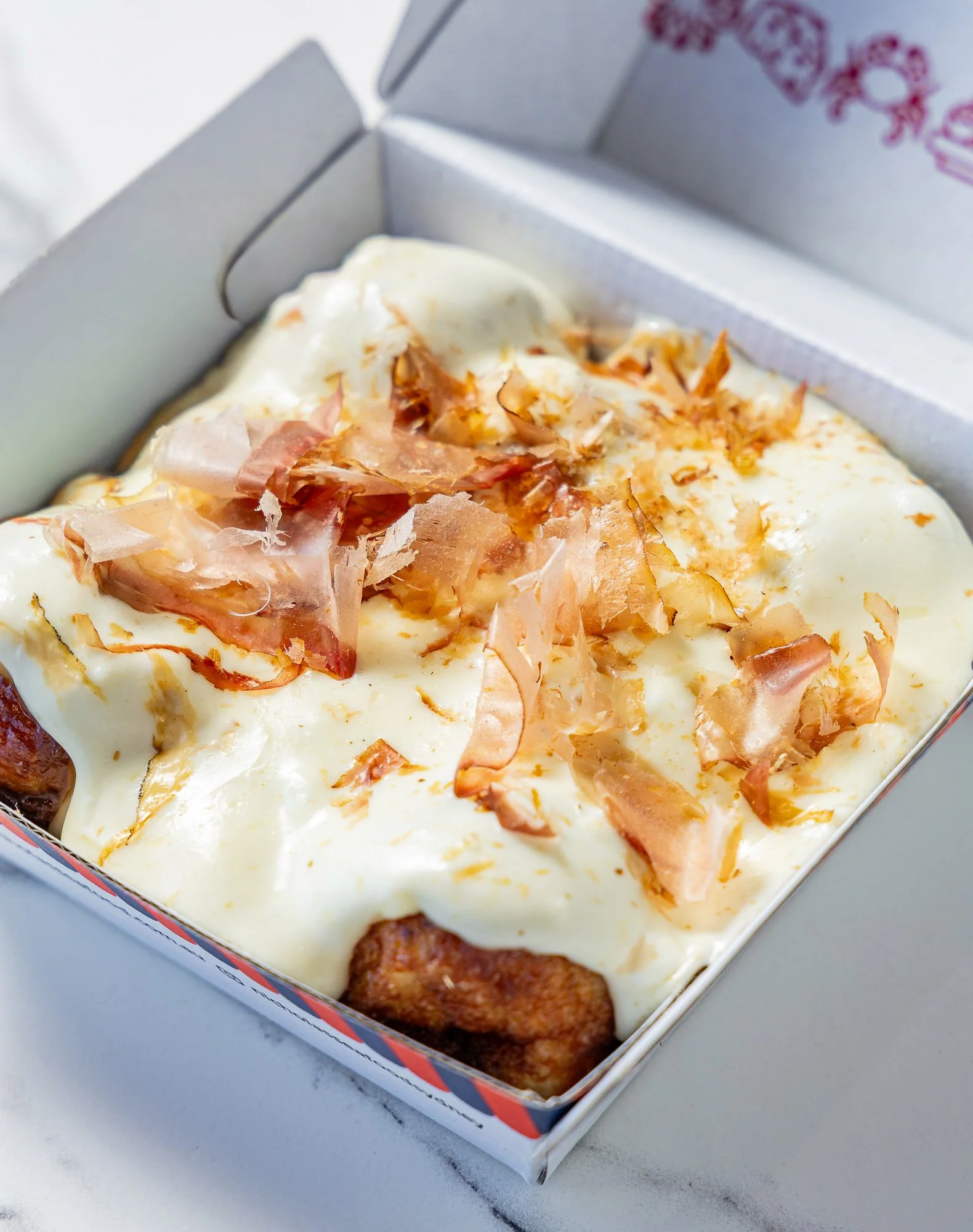 Close-up of a frozen meatloaf topped with cheese, cooked ham, and bacon pieces inside a cardboard box on a white surface.
