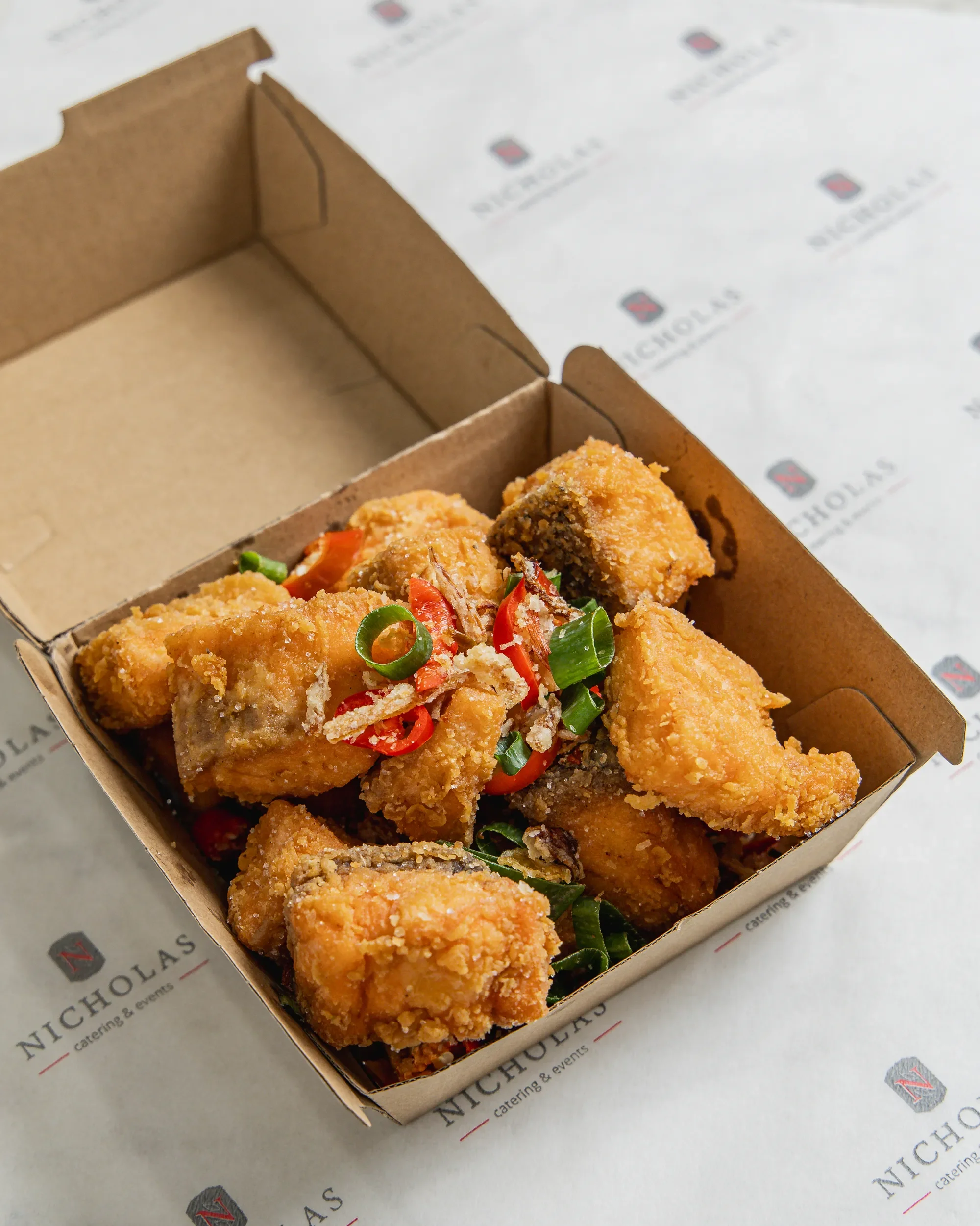 Close-up of a brown cardboard takeout box filled with fried chicken pieces and garnished with sliced red and green peppers on branded paper.
