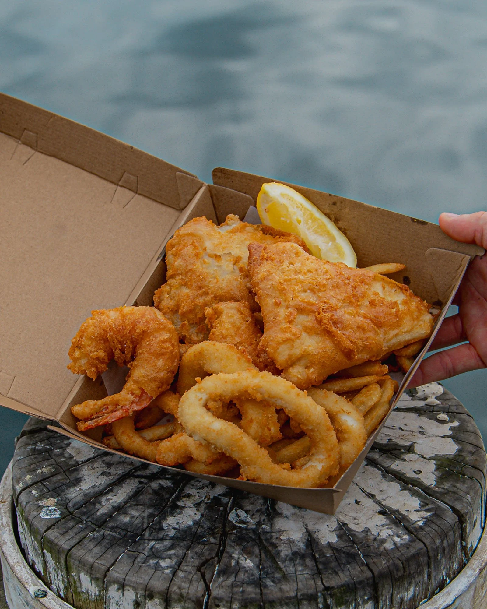 A cardboard takeout box contains fried fish fillets, so fried shrimp, fried onion rings, and a lemon wedge, all placed on a weathered tree stump by water.