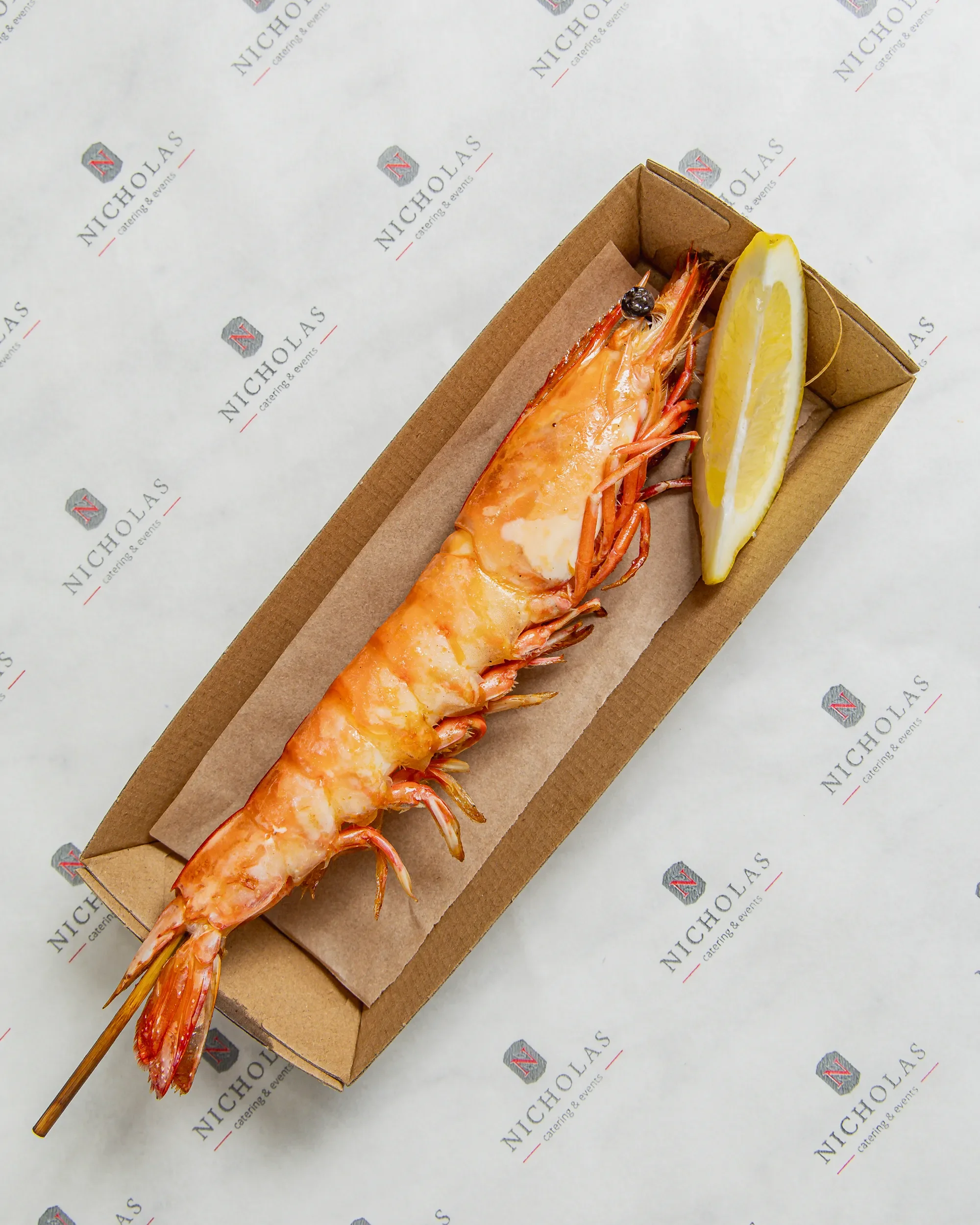 A cooked lobster tail with a lemon wedge on a paper-lined tray, on top of branded paper with 'NICHOLAS catering & events' printing.