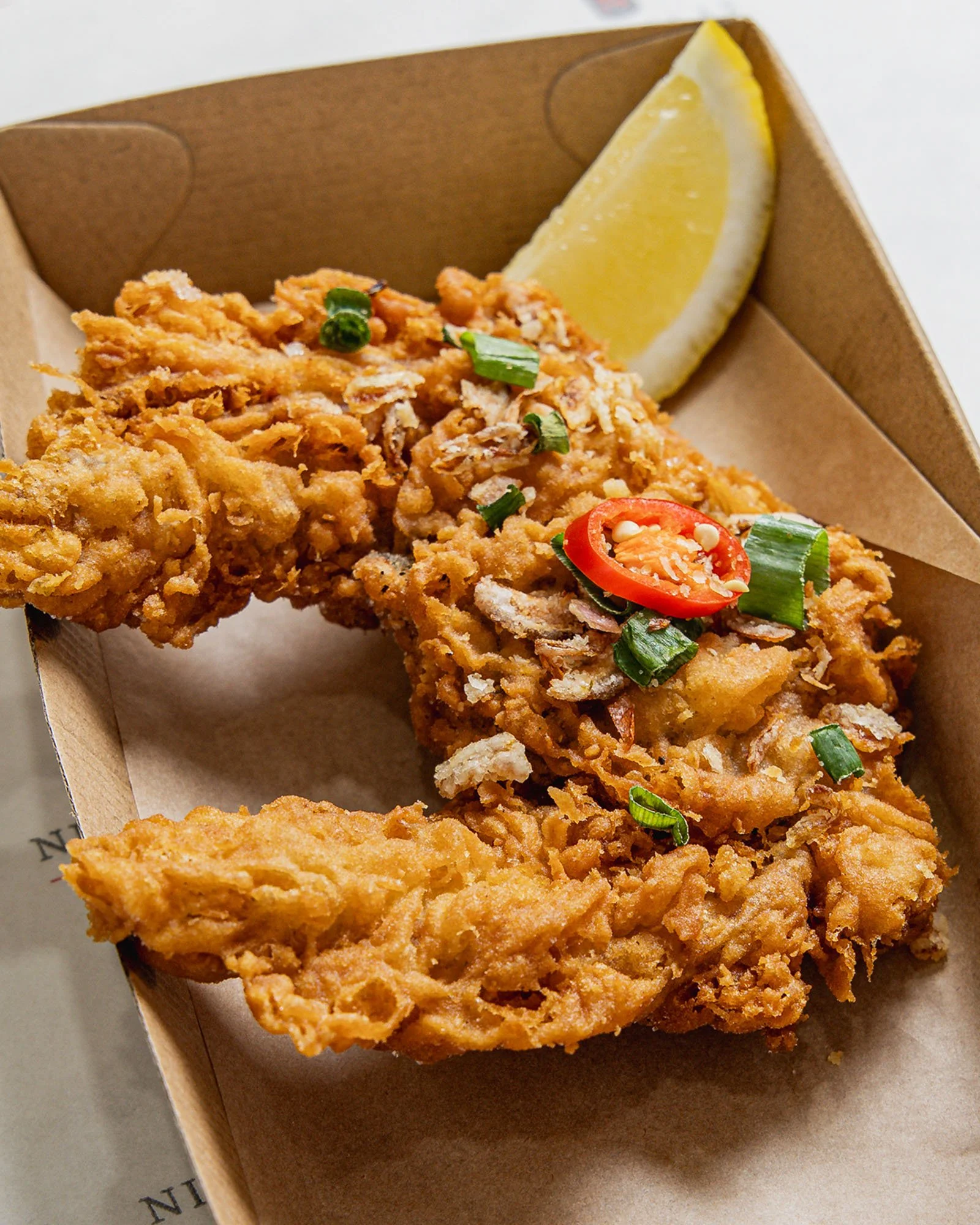 A cardboard takeout box containing fried chicken strips garnished with chopped green onions and sliced red chili, alongside a lemon wedge.