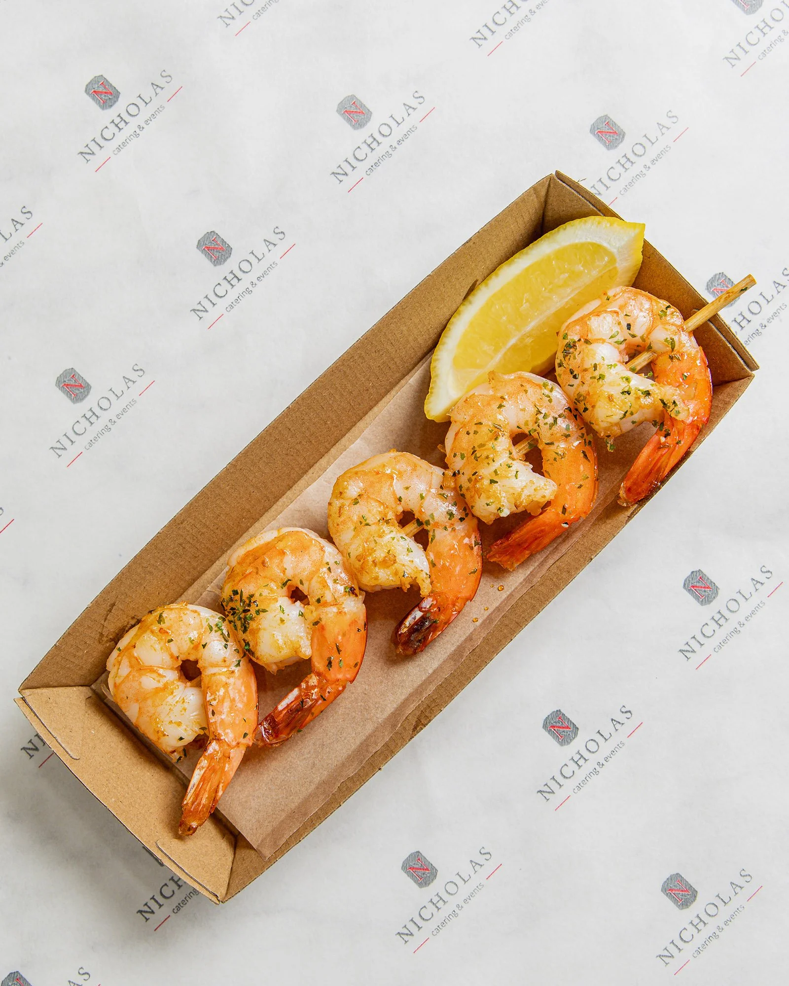 A cardboard serving tray with cooked shrimp garnished with herbs, lemon wedges, and a small wooden skewer, on printed paper with a logo and text.