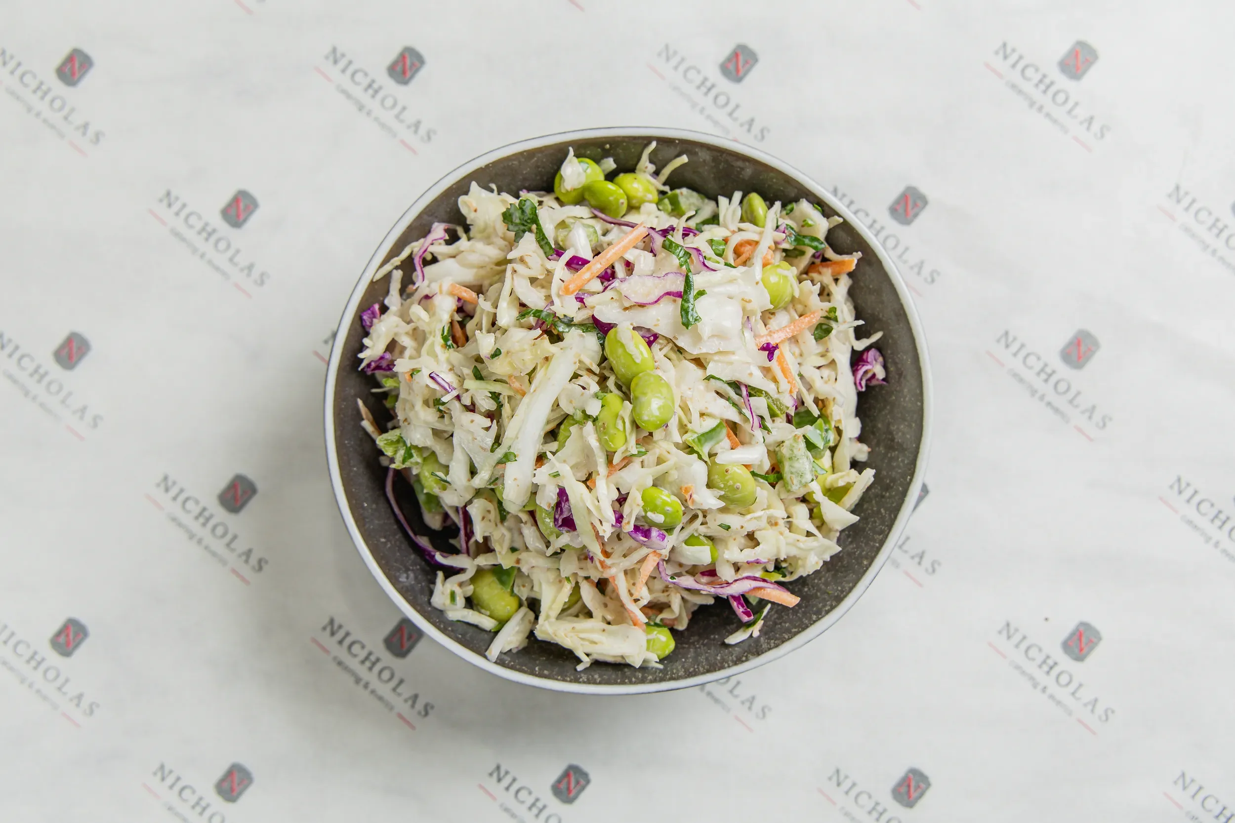 A bowl of coleslaw salad with shredded cabbage, carrots, green peas, and chopped herbs, placed on a white surface with a repeated 'Nicholas' logo pattern.