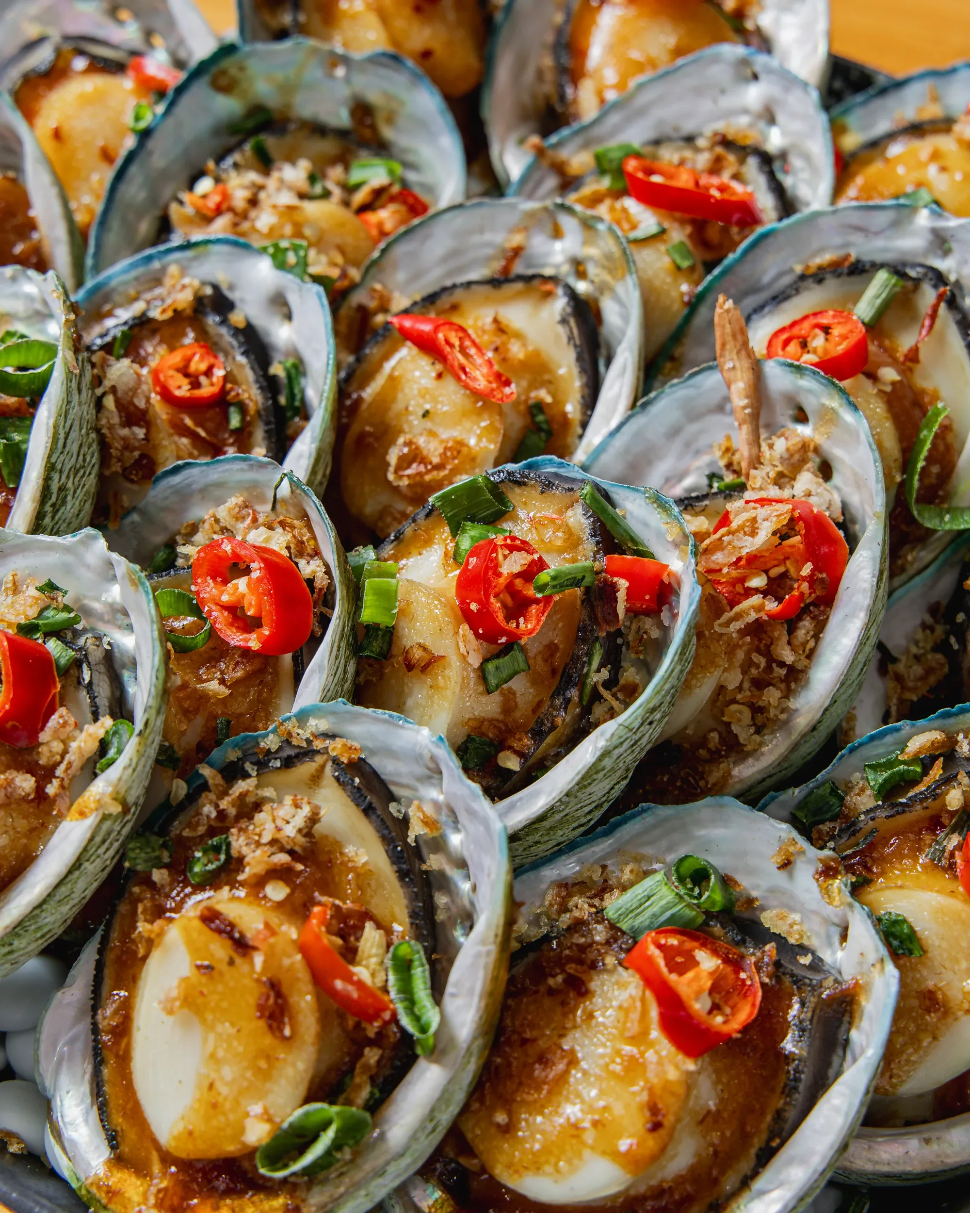 Close-up of baked oysters on the half shell topped with chopped chili peppers, green onions, and breadcrumb crumbs.