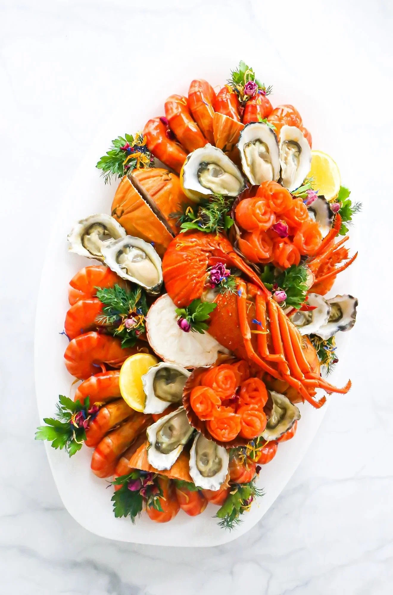A seafood platter featuring lobster, oysters on the half shell, salmon, shrimp, and garnished with fresh herbs and lemon wedges, arranged on a white platter.