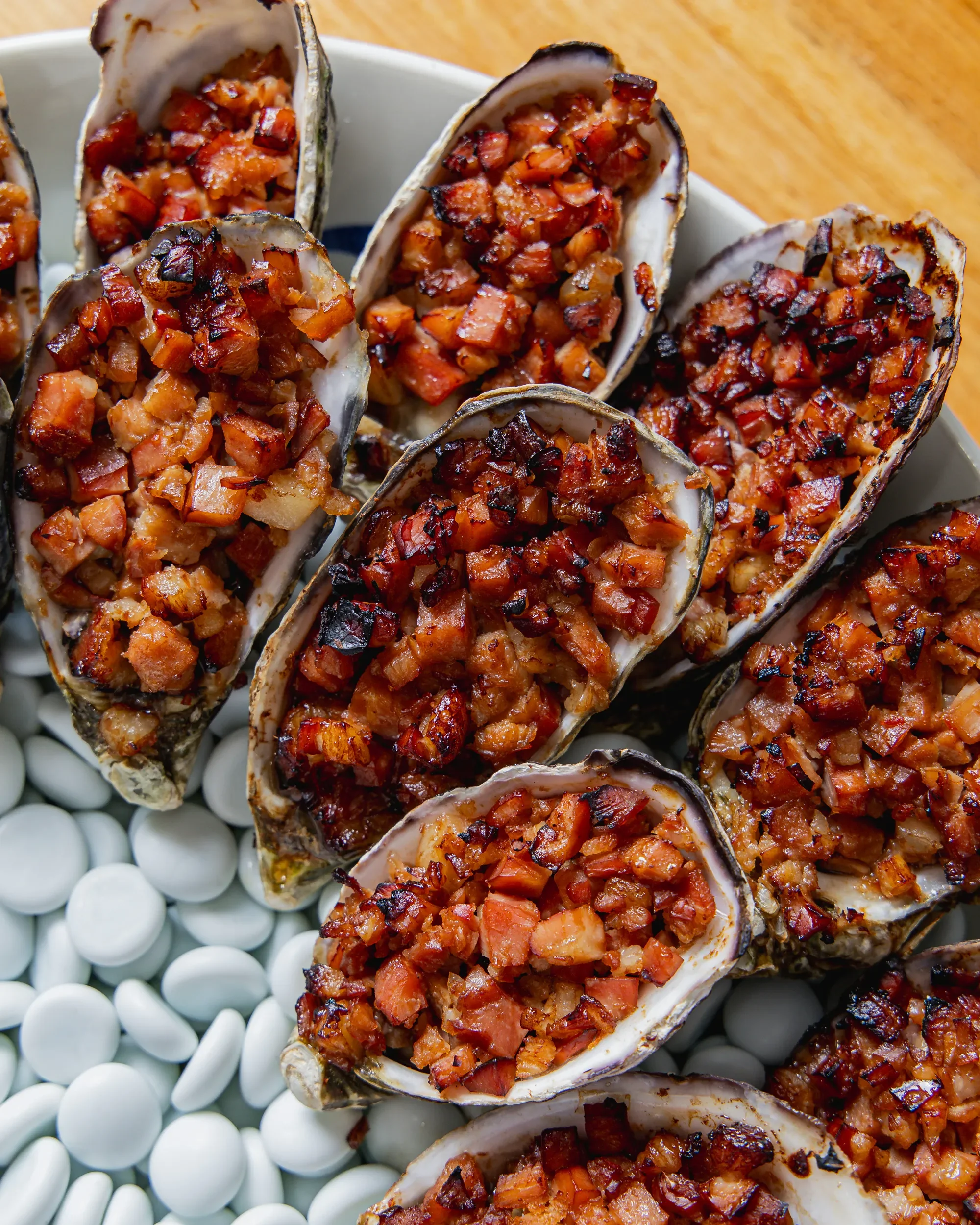 Close-up of baked oysters topped with a mixture of diced bacon and vegetables, on a white plate. A bed of small white round objects is partially visible below the oysters.