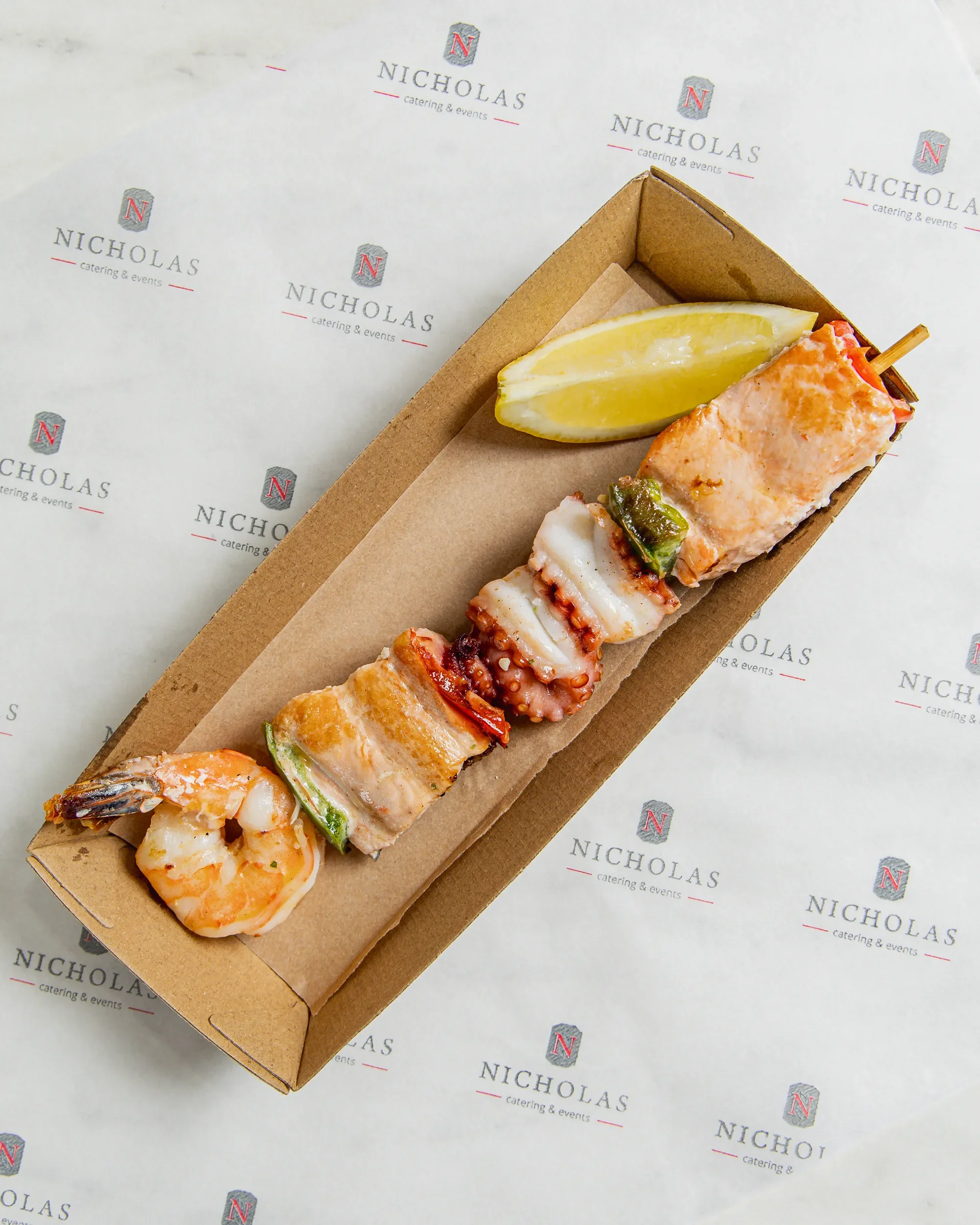 A cardboard tray with grilled seafood skewers, lemon wedges, and a slice of lemon on specialized catering paper with 'Nicholas catering & events' logo repeated on it.