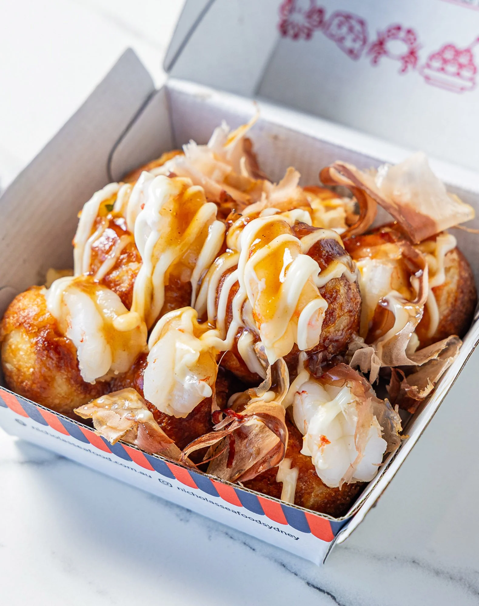 Close-up of a box of fried chicken topped with mayonnaise, drizzled sauce, shredded cheese, and shredded meat, on a white marble surface.