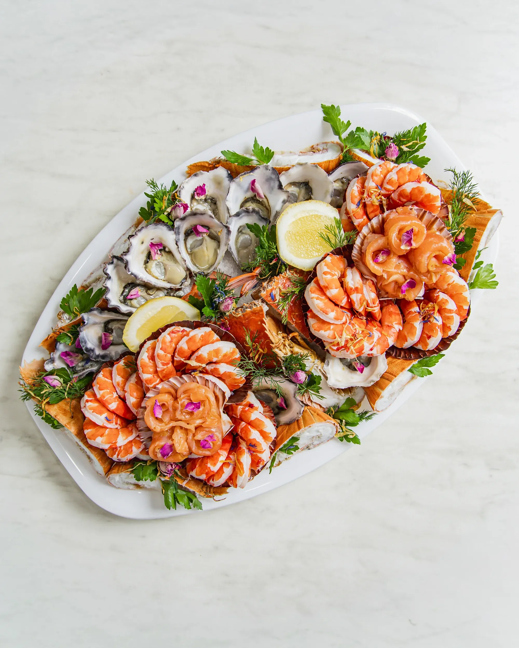 A white plate with fresh oysters on the half shell, garnished with lemon wedges and green herbs, arranged decoratively with small pink flowers.