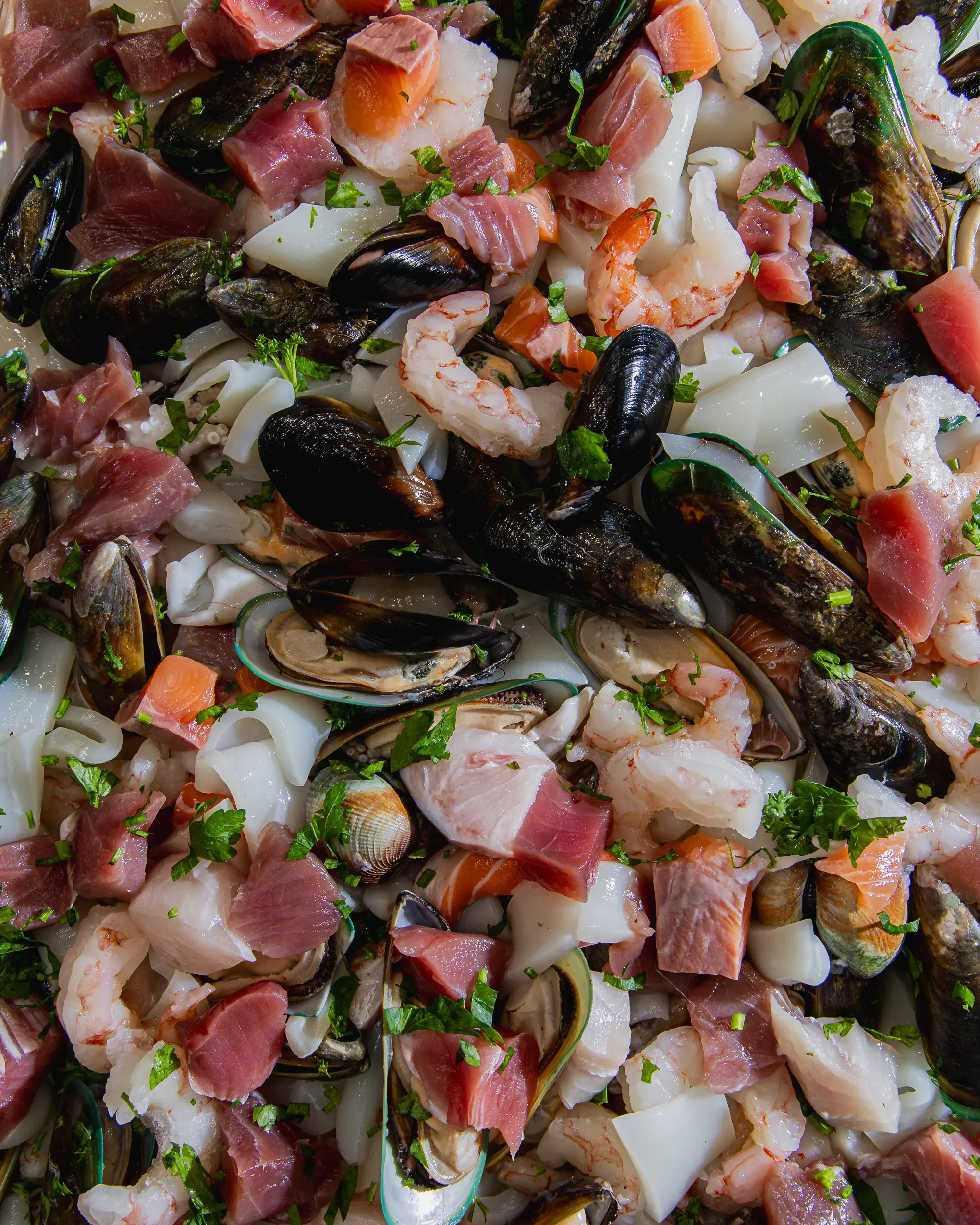 Close-up of a variety of fresh seafood including clams, mussels, shrimp, salmon, and pieces of white fish sprinkled with chopped herbs.