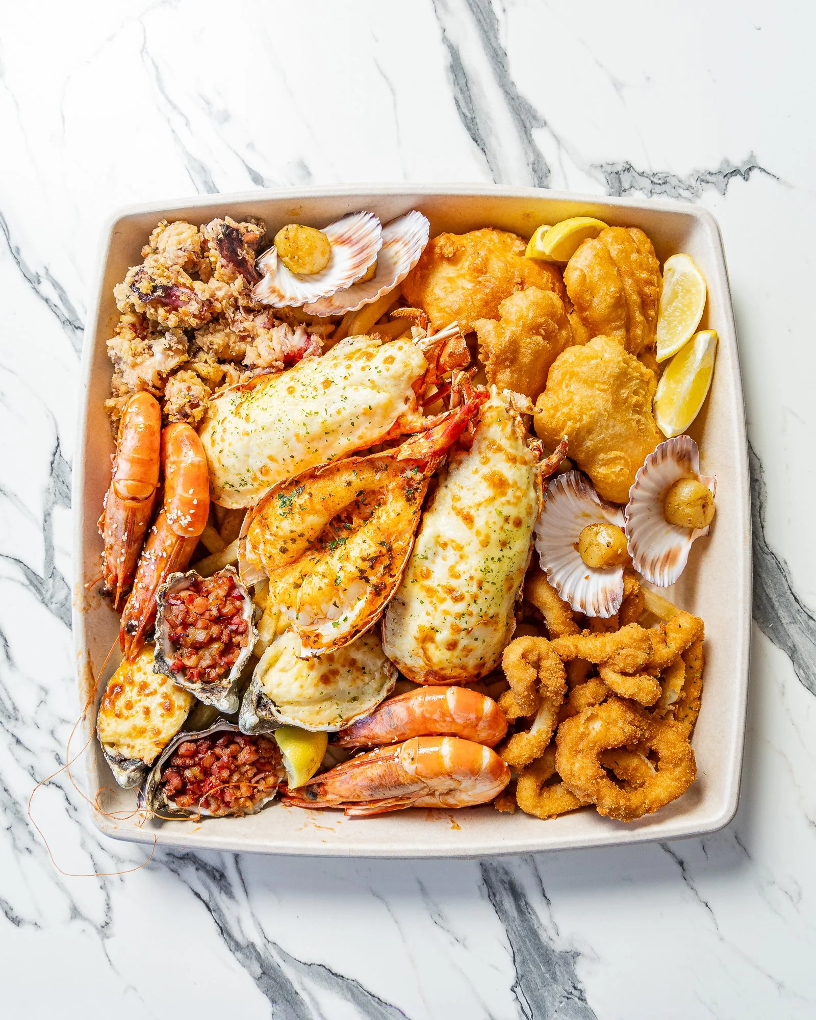 A takeout container filled with fried seafood, shrimp, fish, and crab cakes, garnished with lemon wedges and surrounded by shells and seafood.
