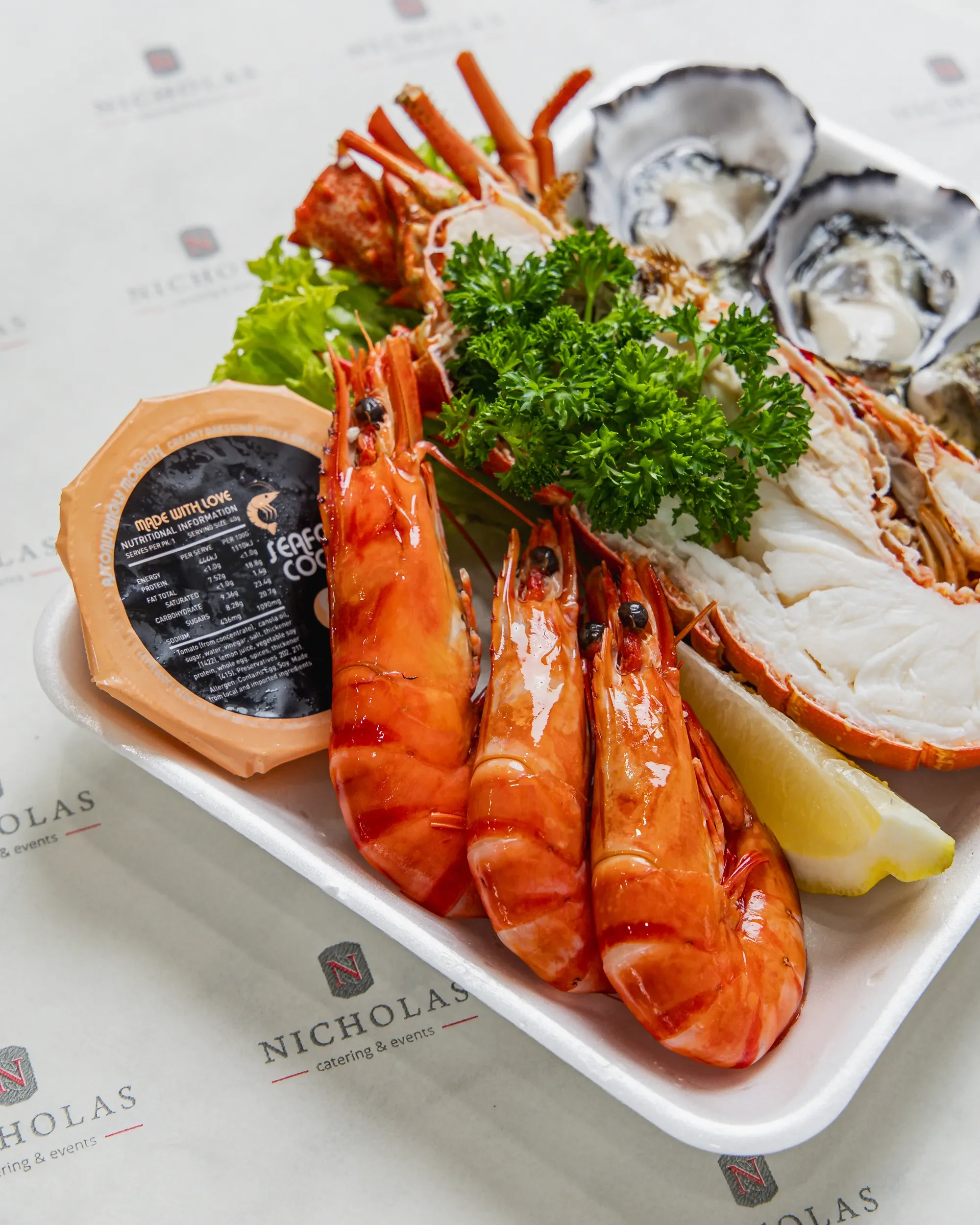 A seafood platter with lobster tail, oysters on the half shell, lemon wedge, greenery, and a packet of cocktail sauce, presented on a styrofoam tray with branding printed on the paper underneath.