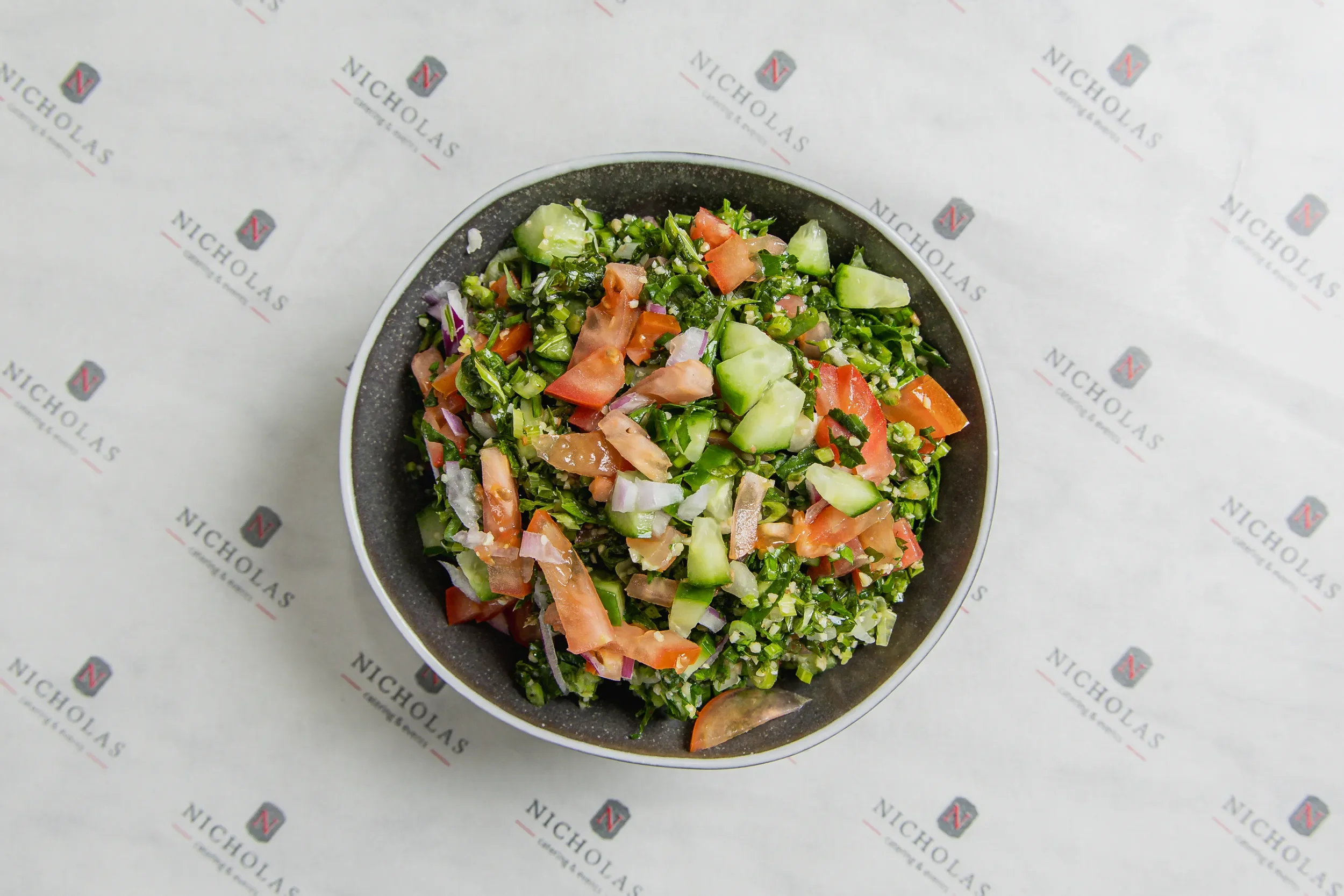 A bowl of chopped salad with cucumbers, tomatoes, onions, and greens on a paper liner with 'Nicholas' logo.