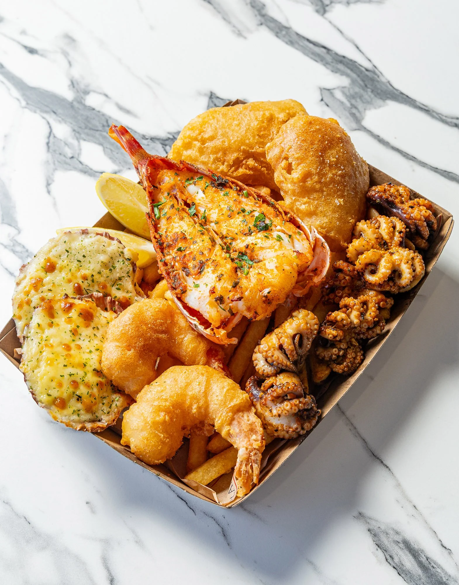 A box of seafood with fried shrimp, battered fish, scalloped potatoes, fried octopus, and French fries on a marble surface.