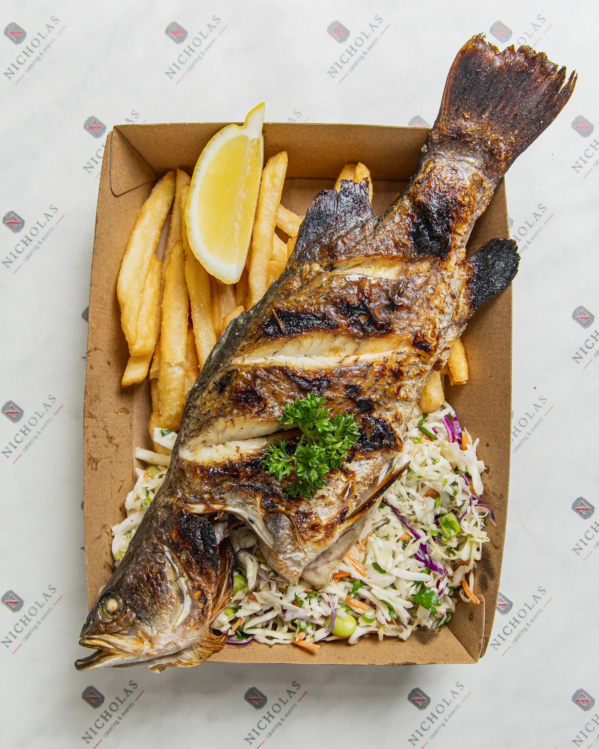 A grilled fish served with French fries, a lemon wedge, and coleslaw, presented on a brown tray with a background featuring the Nicholaz branding.