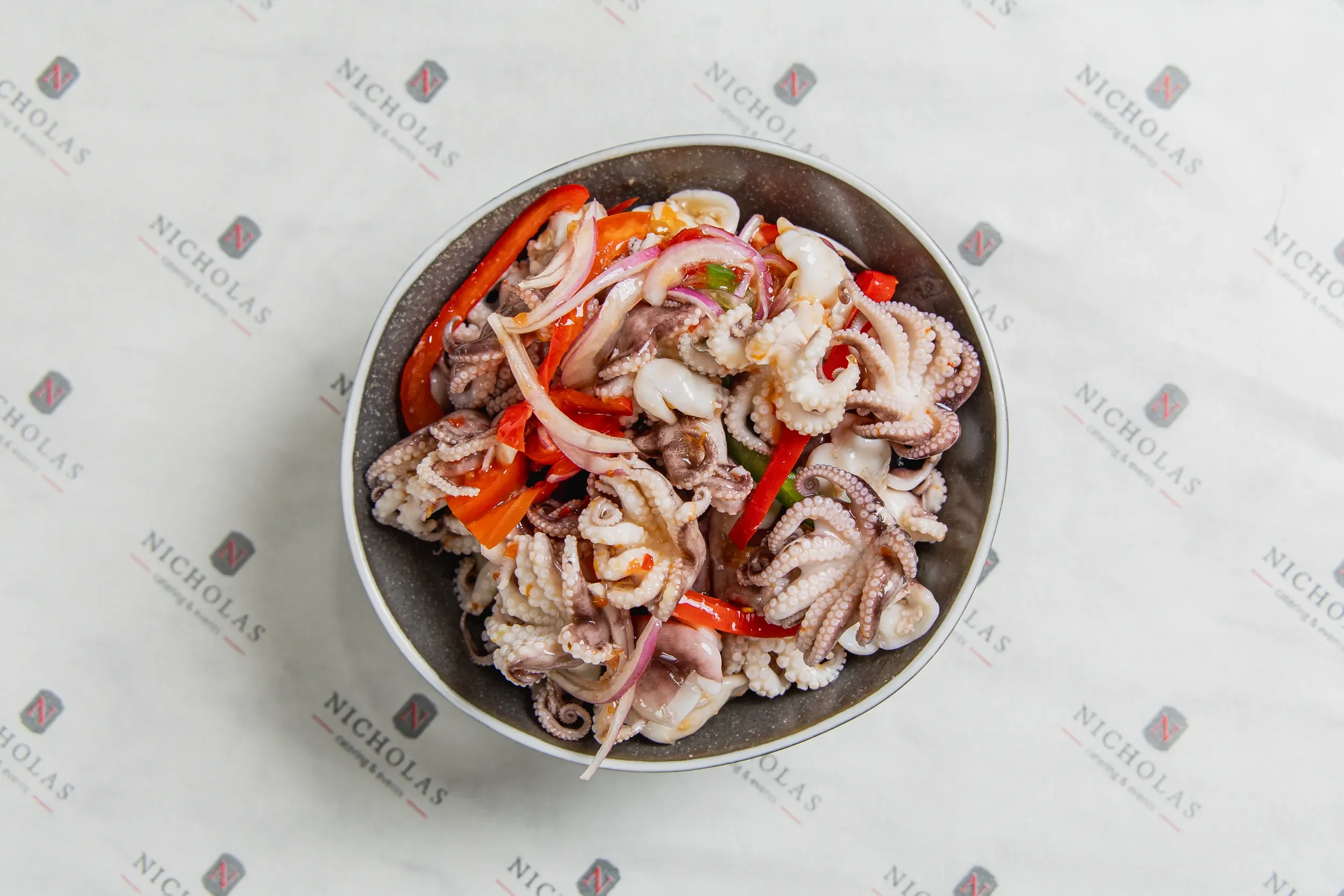 A bowl filled with mixed seafood including octopus tentacles, sliced red bell peppers, and other seafood pieces on white paper with 'Nicholas' branding.