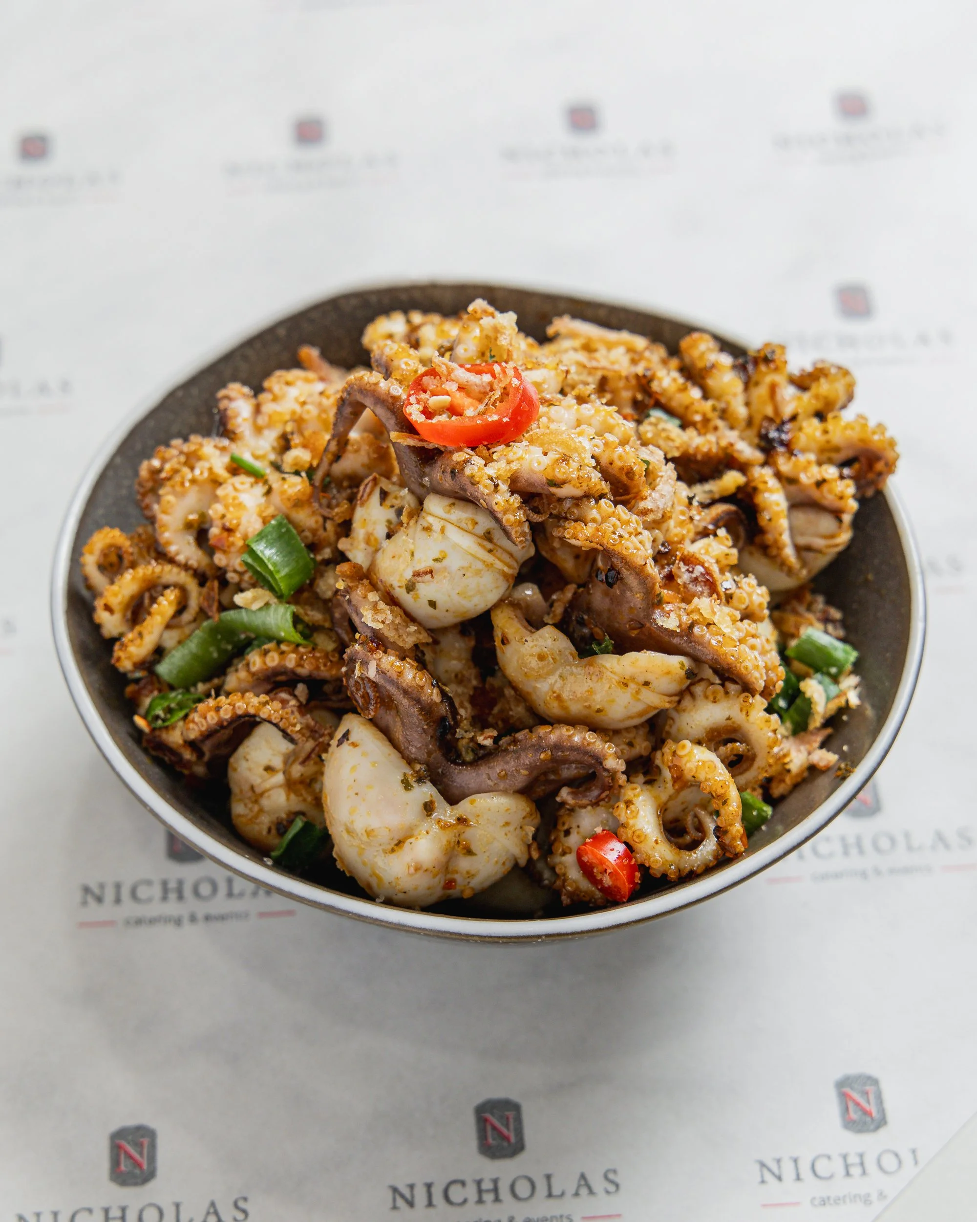 A bowl of grilled seafood including squid and octopus, garnished with sliced green onions and red chili peppers, on a paper with "Nicholas catering & events" branding.