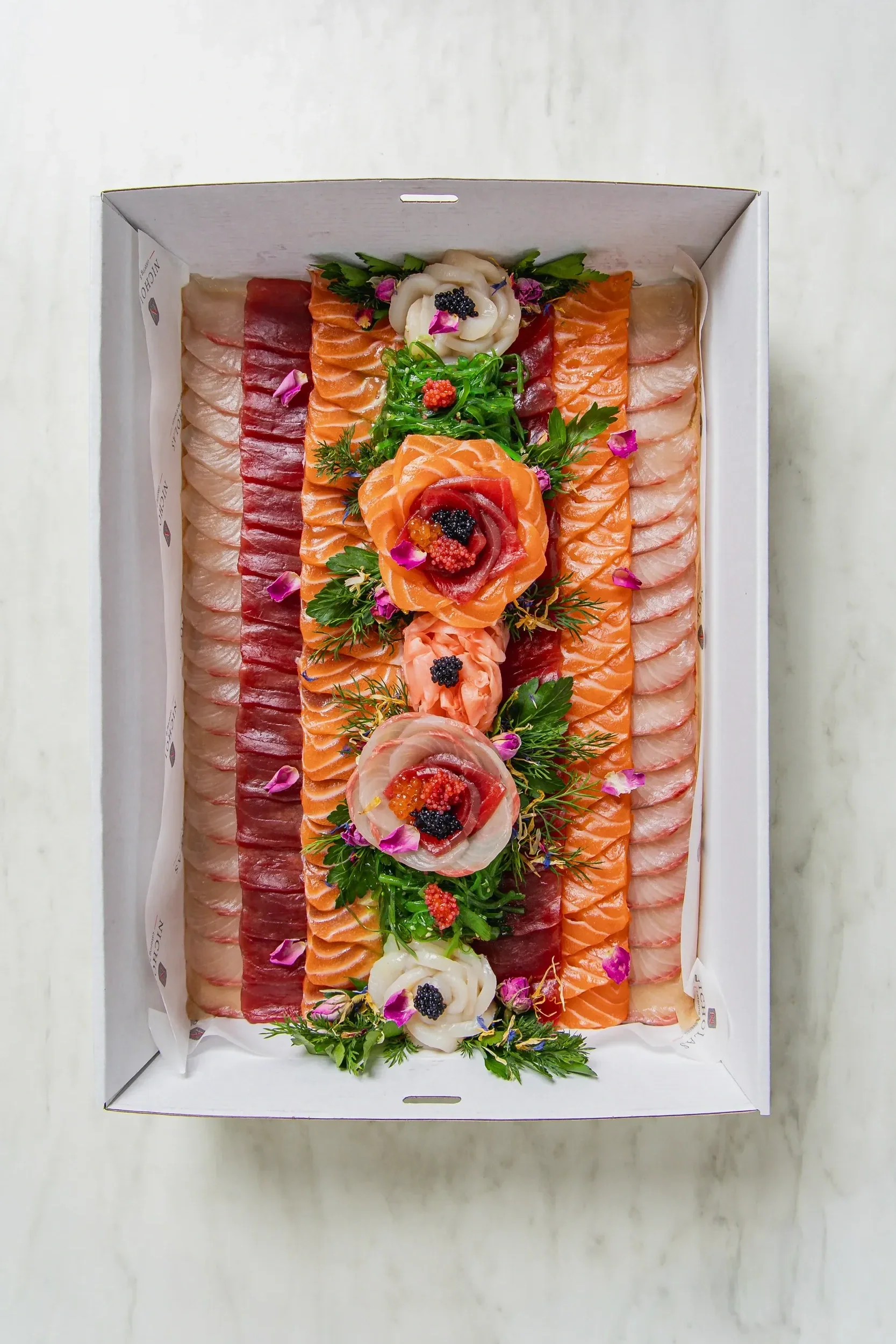 A white box filled with assorted sliced raw fish, including salmon, tuna, and whitefish, garnished with herbs, edible flowers, and small dollops of caviar.
