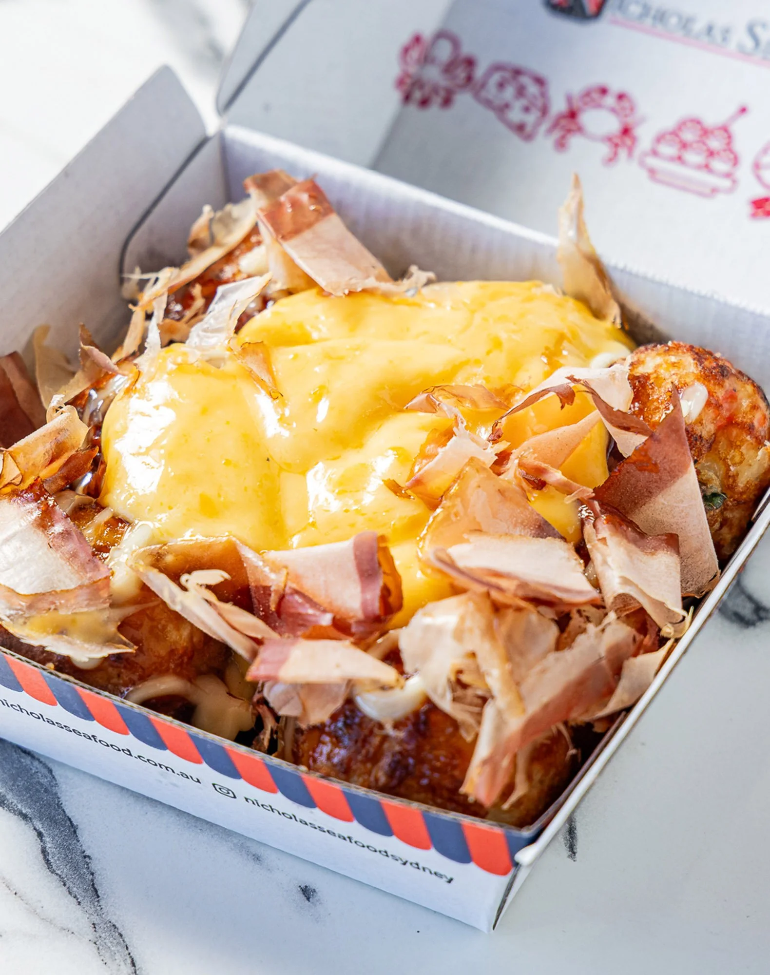 A close-up of a pizza with cheese, ham, and possibly other toppings in an open pizza box on a white surface.