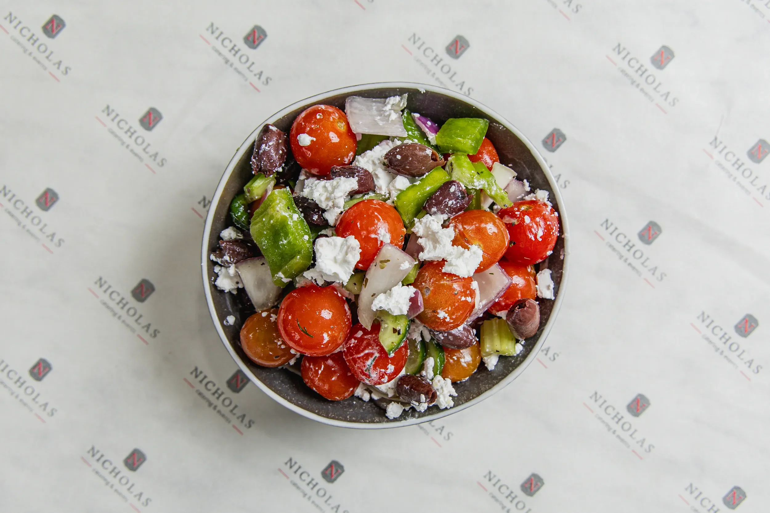 A bowl filled with cherry tomatoes, green bell pepper, onion, olives, feta cheese, and herbs on branded paper background.