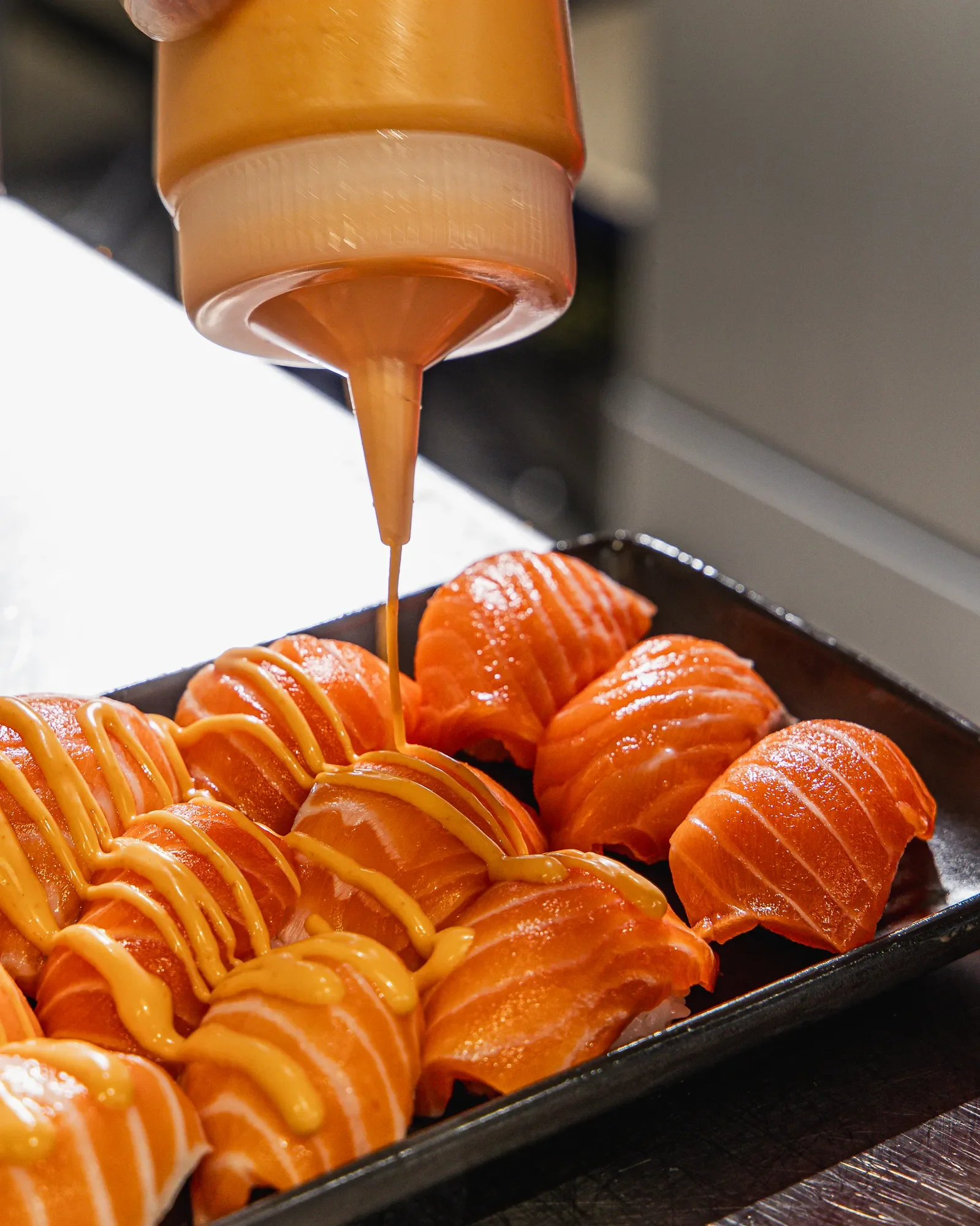 Close-up of fresh, sliced salmon sashimi drizzled with orange sauce and topped with mayonnaise in a black tray.