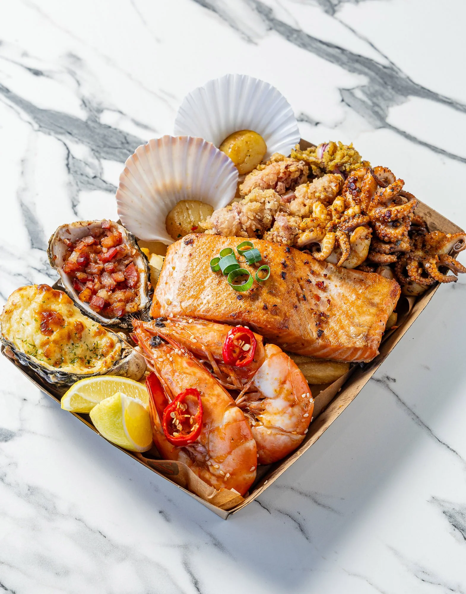 A seafood platter with grilled salmon, shrimp, octopus, stuffed crab, roasted potatoes, lemon wedges, and garnishes served on a rectangular tray on a marble surface.