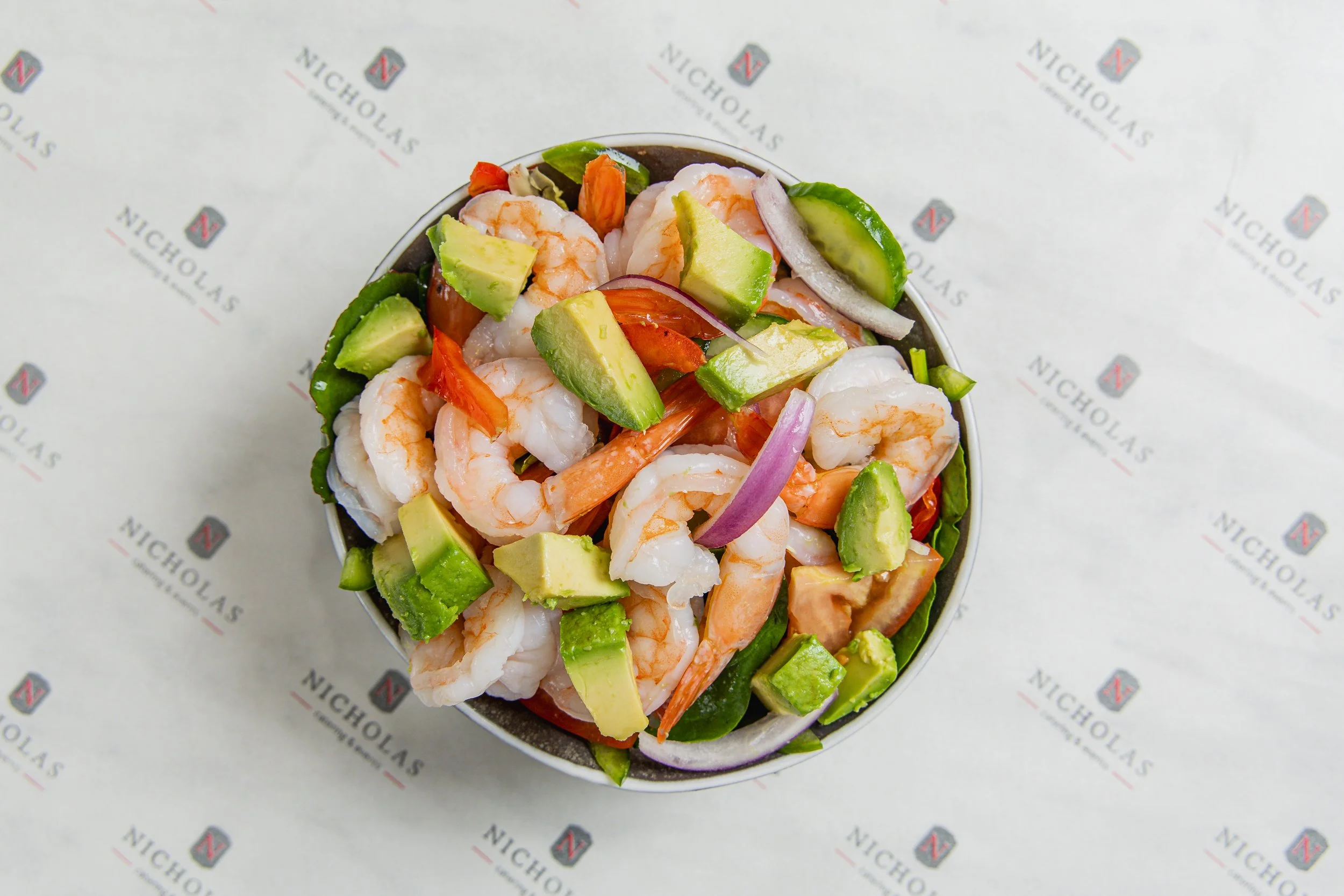Close-up of a seafood salad with shrimps, avocado, sliced onions, and tomatoes in a bowl on branded paper.