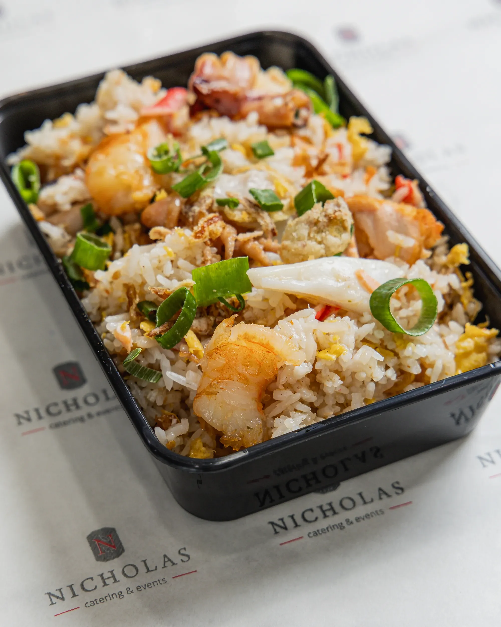 Bento box with rice, shrimp, chicken, eggs, green onions, and vegetables placed on branded paper with 'Nicholas catering & events'.