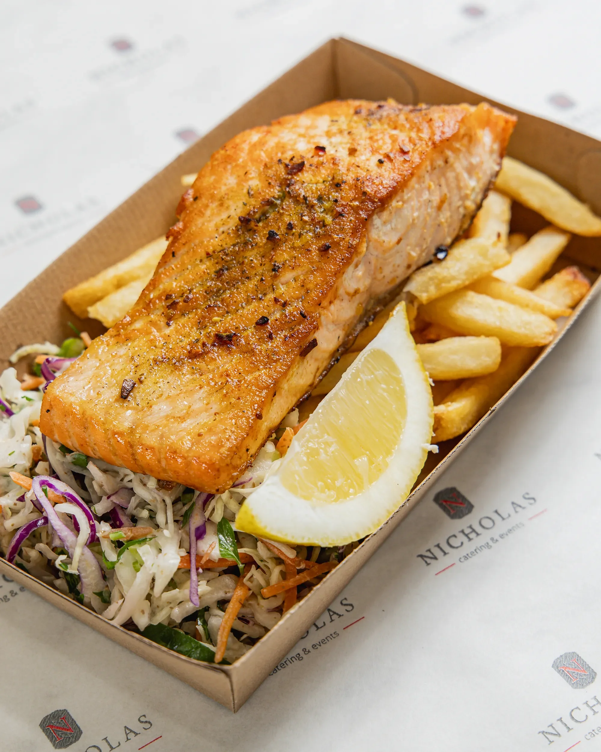 Grilled salmon fillet with crispy skin served with lemon wedges, and a side of coleslaw and French fries in a cardboard tray on a paper-lined surface.