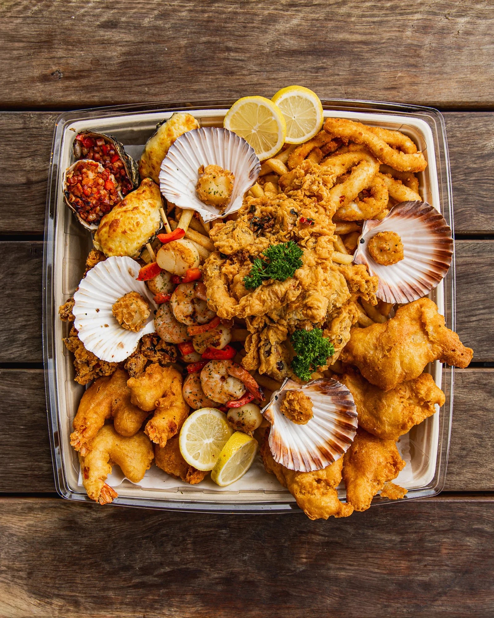 A seafood platter with fried fish, shellfish, lemon slices, and garnishes on a tray placed on a wooden table.