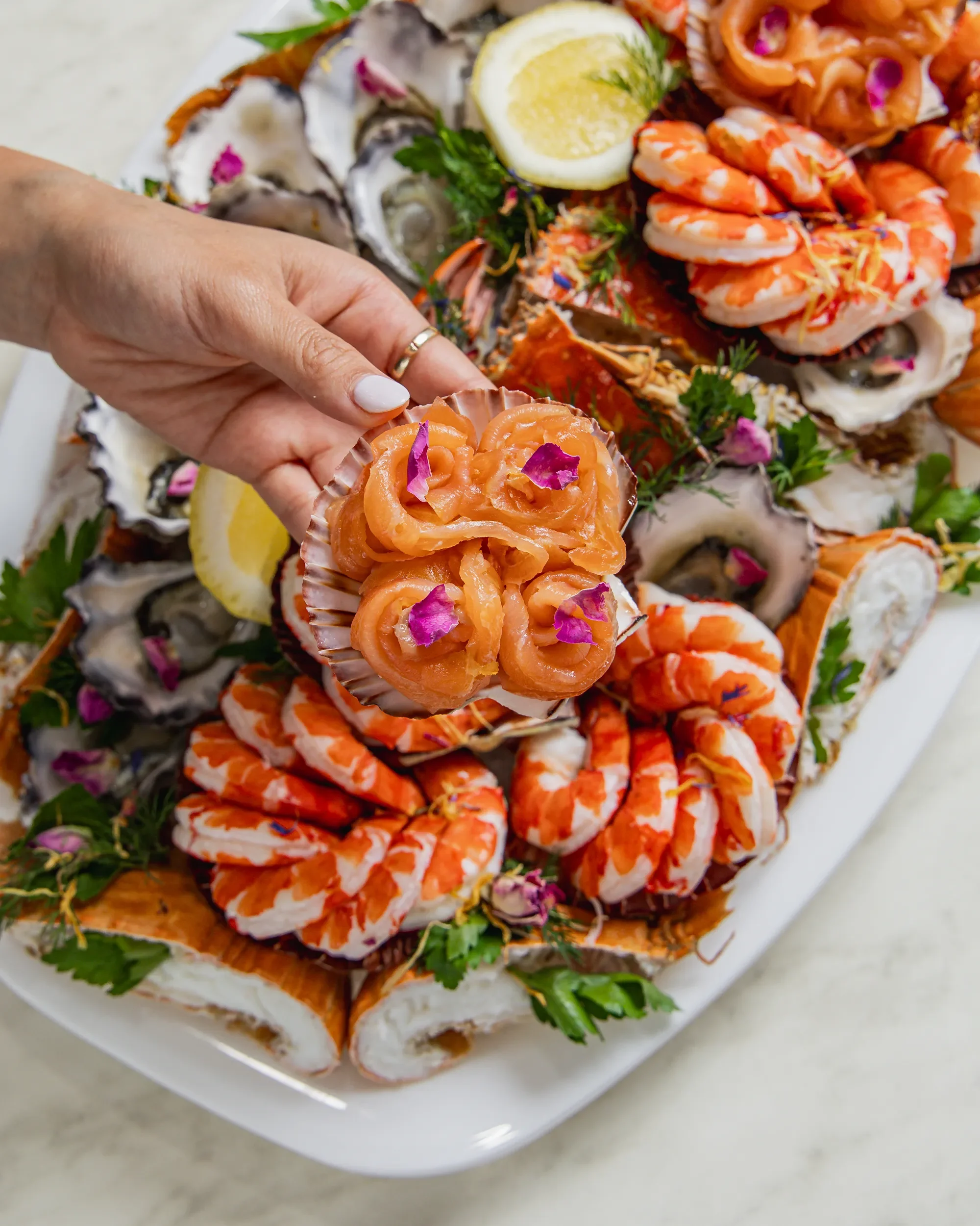 Assorted seafood platter with smoked salmon, shrimp, oysters, lemon slices, and garnishes on a white serving plate.