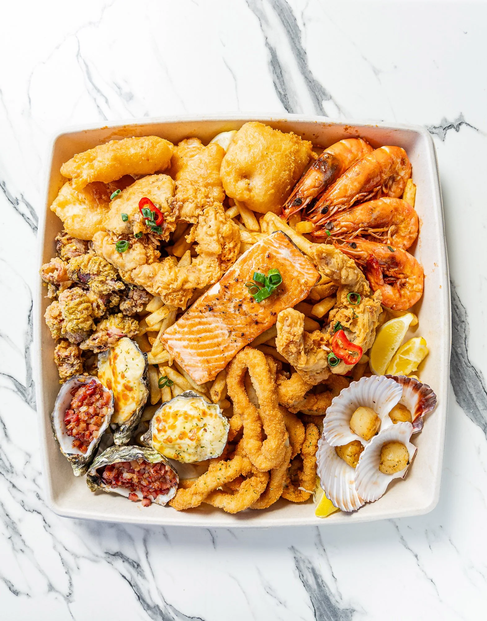 A takeout box filled with fried shrimp, battered fish, scallops, and various seafood dishes on a marble surface.