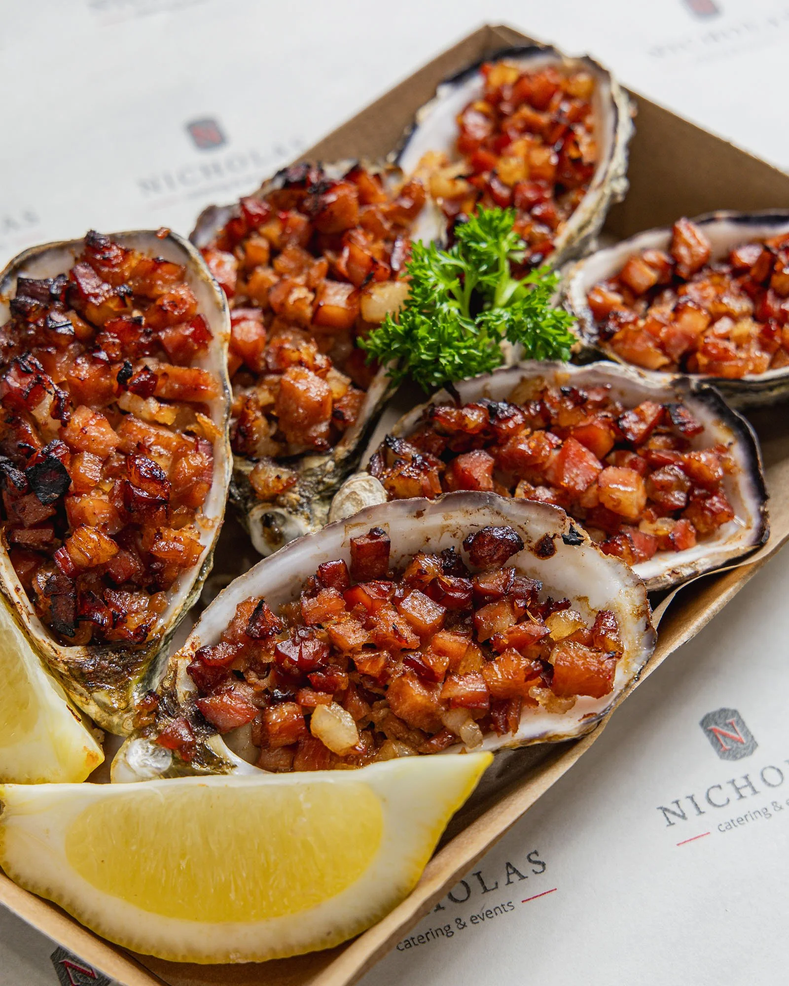 Close-up of baked oysters topped with diced bacon, served with lemon wedges and garnished with parsley.