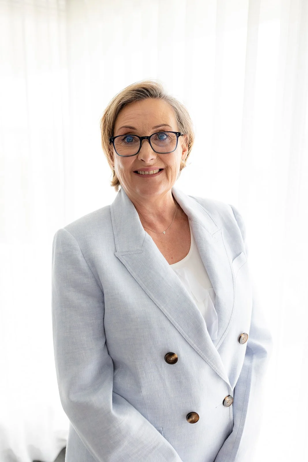 Pamela, looking towards the camera, smiling, with short blonde hair and glasses. She is wearing a light blue blazer with dark buttons and a white top, standing against a bright, white background.