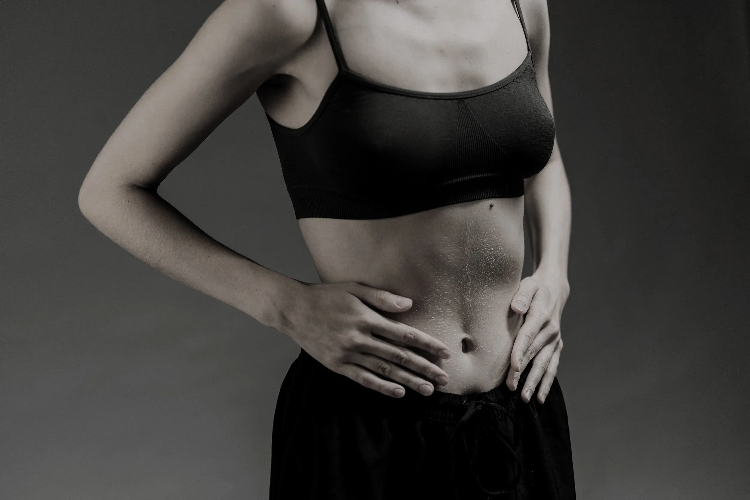 A close-up of a woman's midsection, showing her abdomen with stretch marks, wearing a black sports bra and black shorts against a gray background.