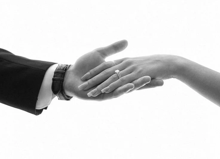 A man in a suit and a woman are holding hands, with the woman's hand showing an engagement ring.